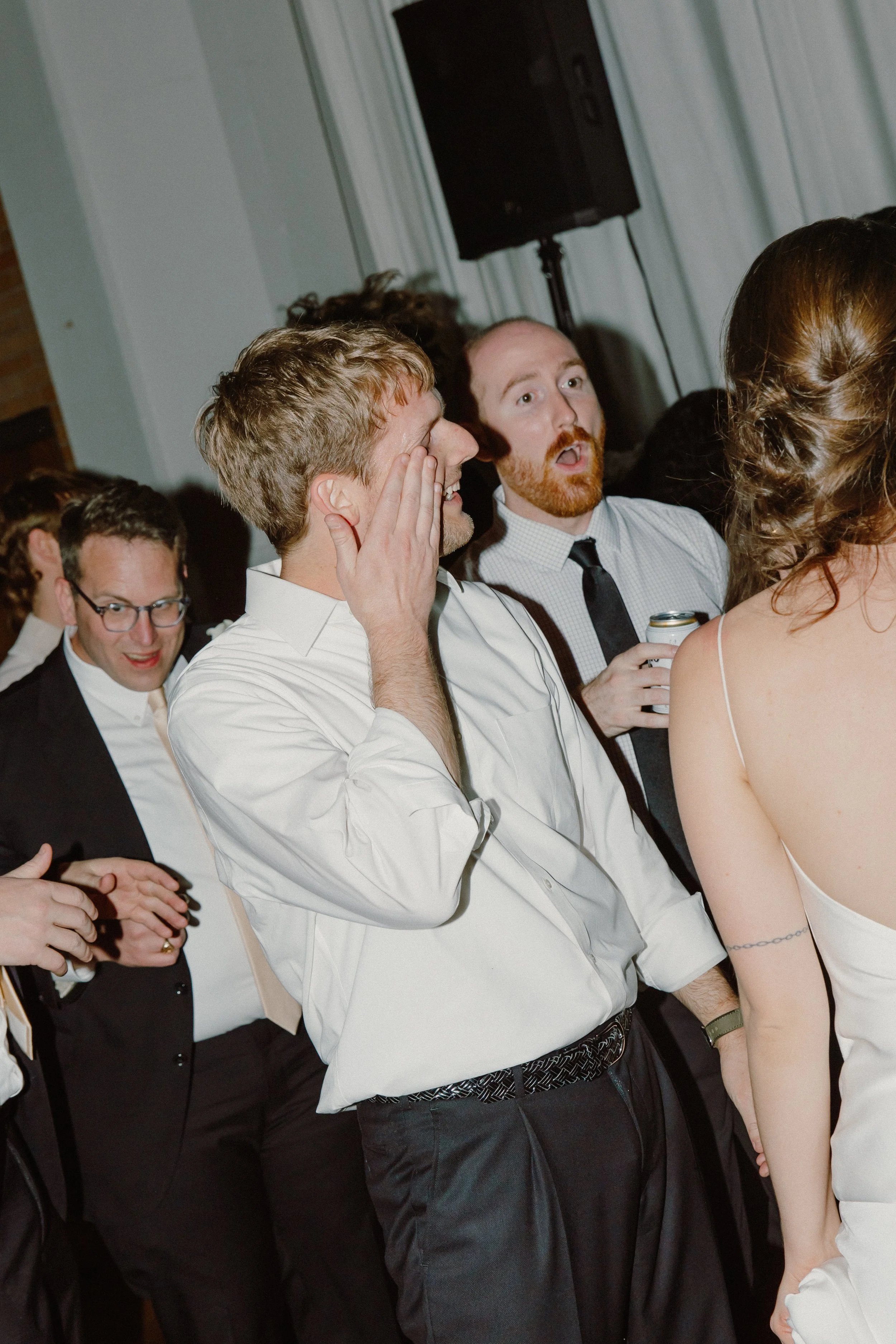 Group of men and women at a party or wedding, some smiling and holding drinks, one man covering his mouth in surprise or laughter.