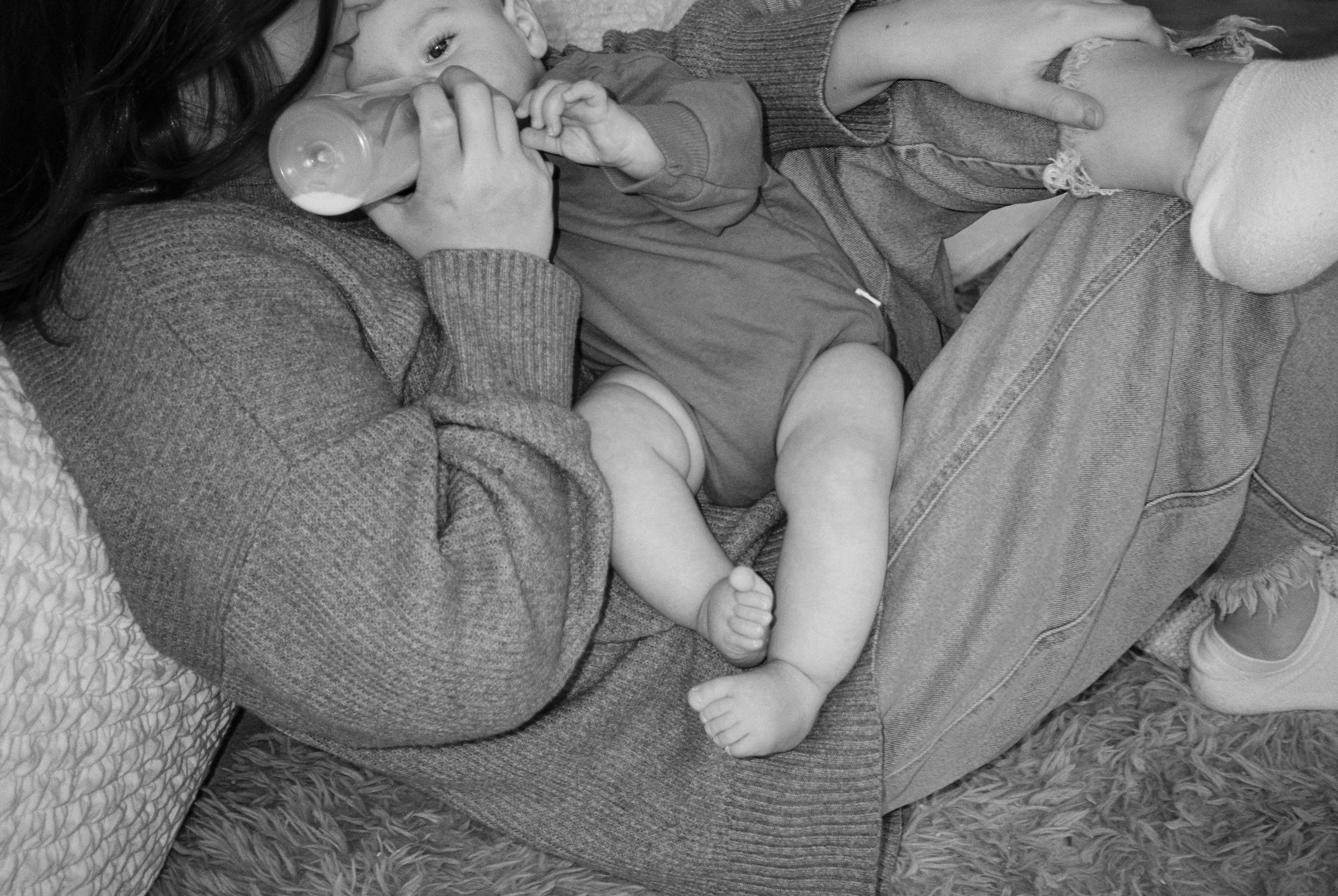 A woman and a young child lying on a bed, with the woman feeding the child with a bottle. The woman wears a sweater, and the child has a onesie and is barefoot.