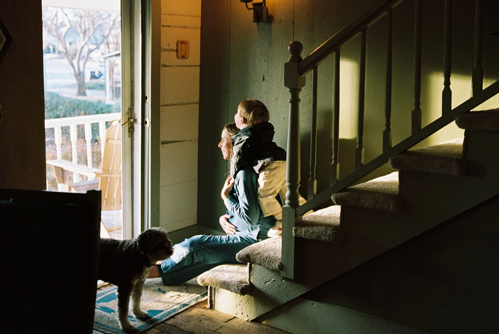 A woman, a child, and a dog sit at the open door of a house, with sunlight shining inside. The woman is sitting on the floor, and the child is on her shoulders. The dog is lying on the floor near the door, and a porch with a chair is visible outside.
