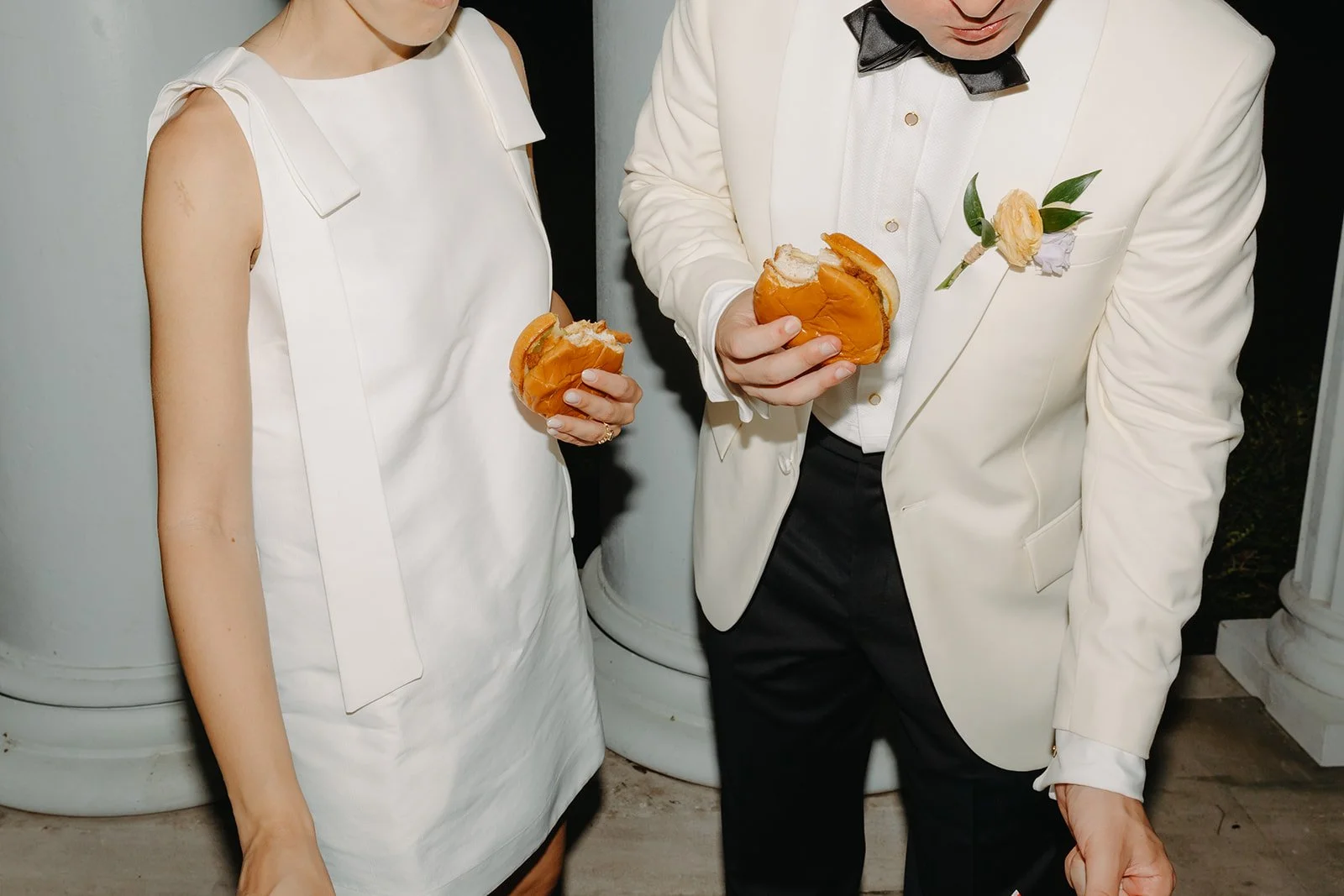 A man and woman dressed formally, holding burgers, at a formal event or wedding reception.