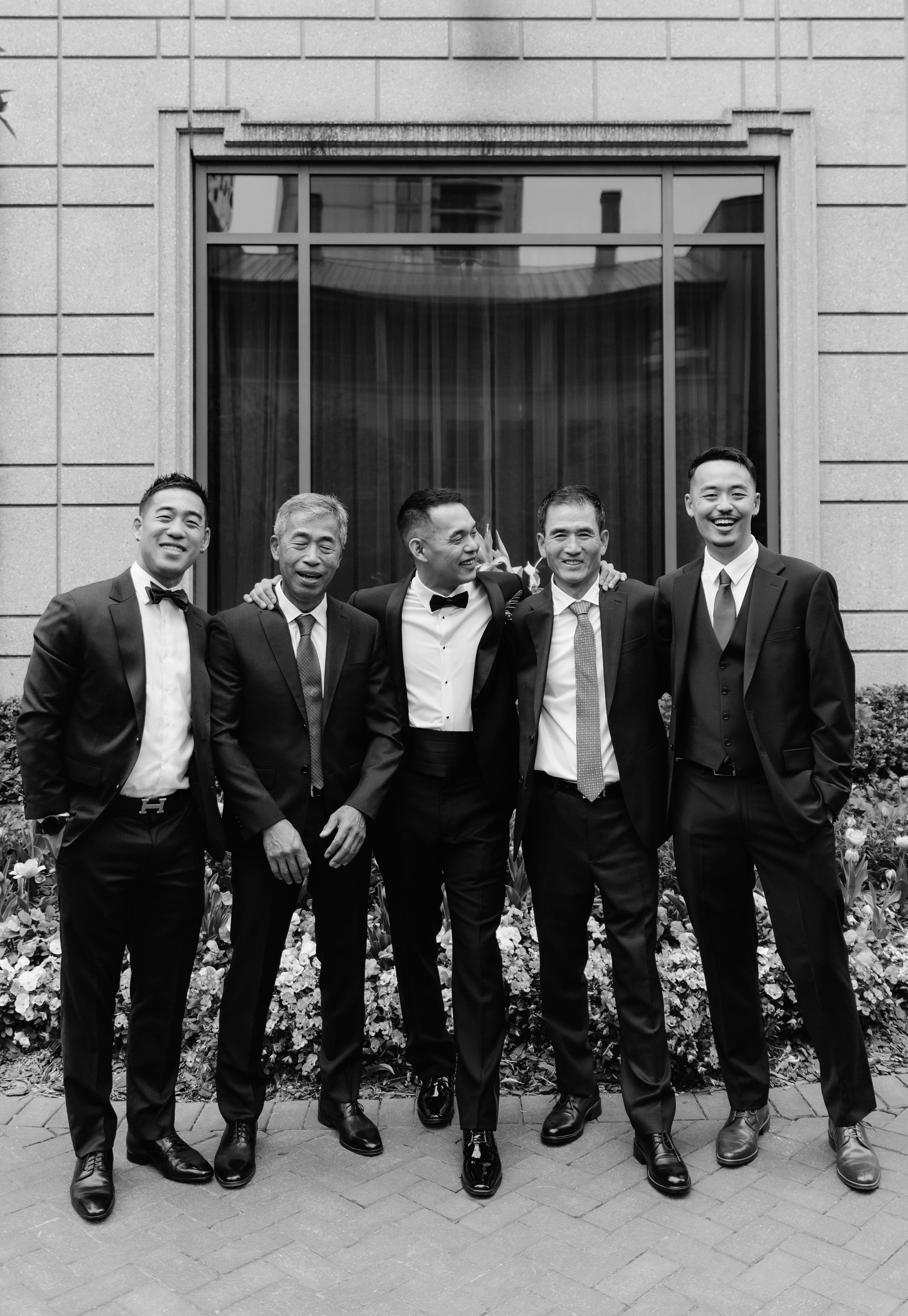A group of five men dressed in tuxedos, standing outside in front of a building with a large window, smiling and enjoying each other's company.