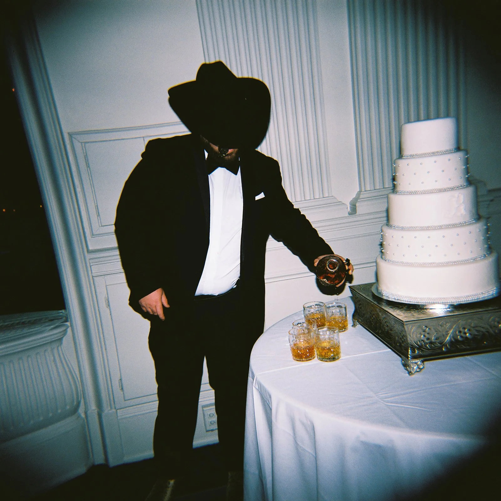 A man wearing a black tuxedo and bow tie, with a fedora hat, pouring a drink at a wedding reception. There is a large white-tiered wedding cake on a silver stand next to him, and several glasses of whiskey or similar drinks on the table.