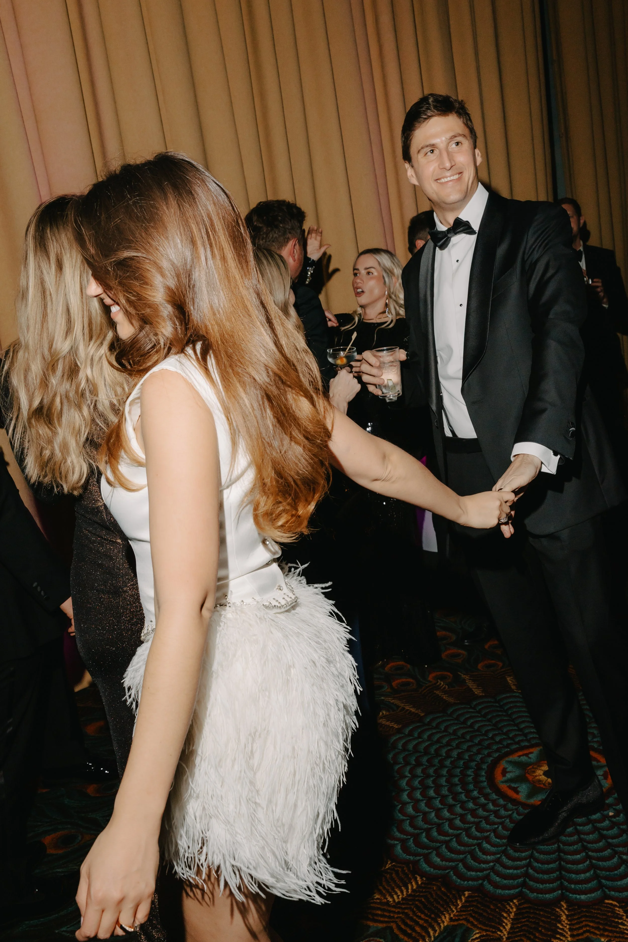 A group of elegantly dressed people dancing and socializing at a formal event, with a focus on a man in a tuxedo holding hands with a woman in a white dress with feather details.