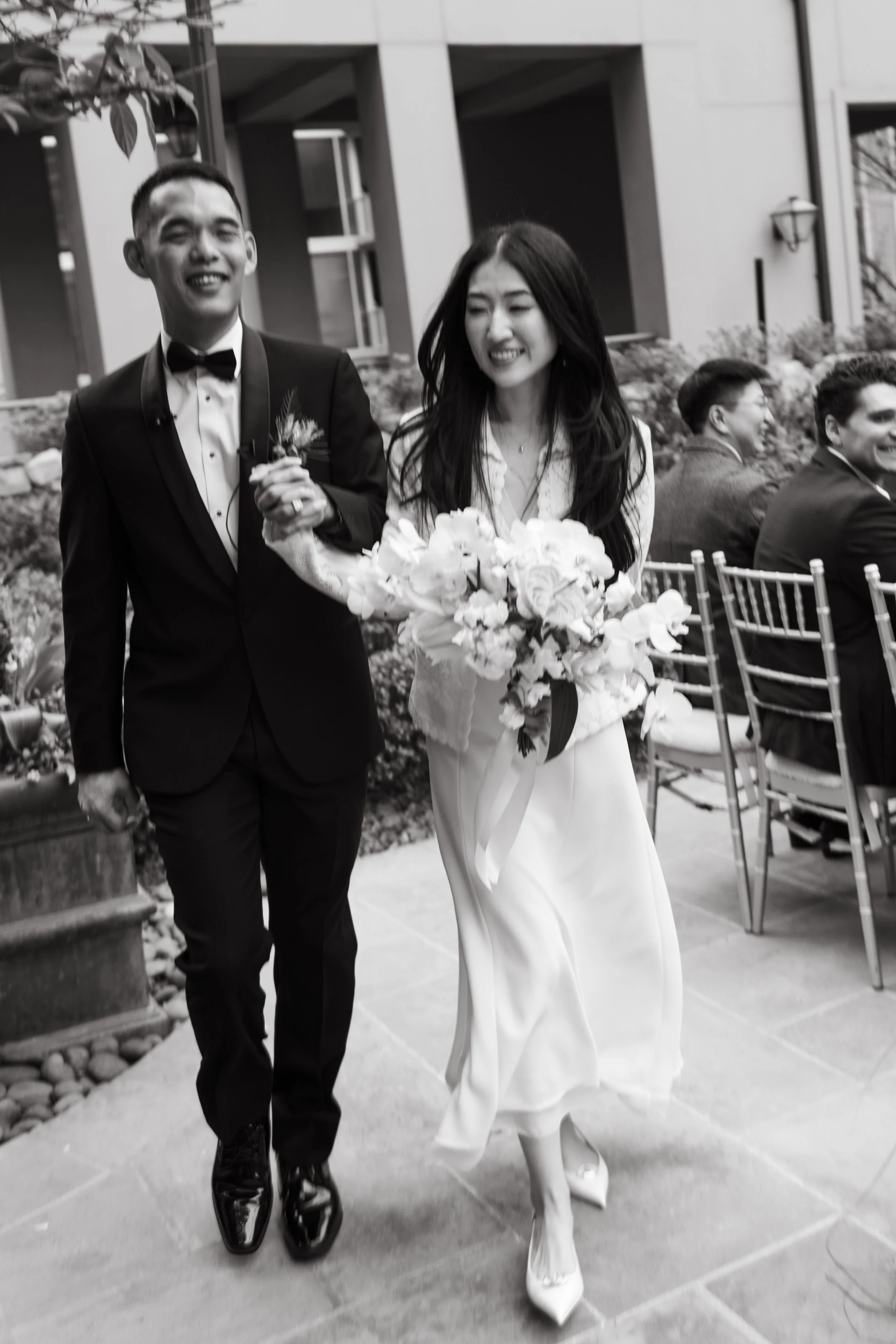 A newlywed couple, a man in a tuxedo and a woman in a white dress, walking arm in arm with smiles, during their wedding celebration.