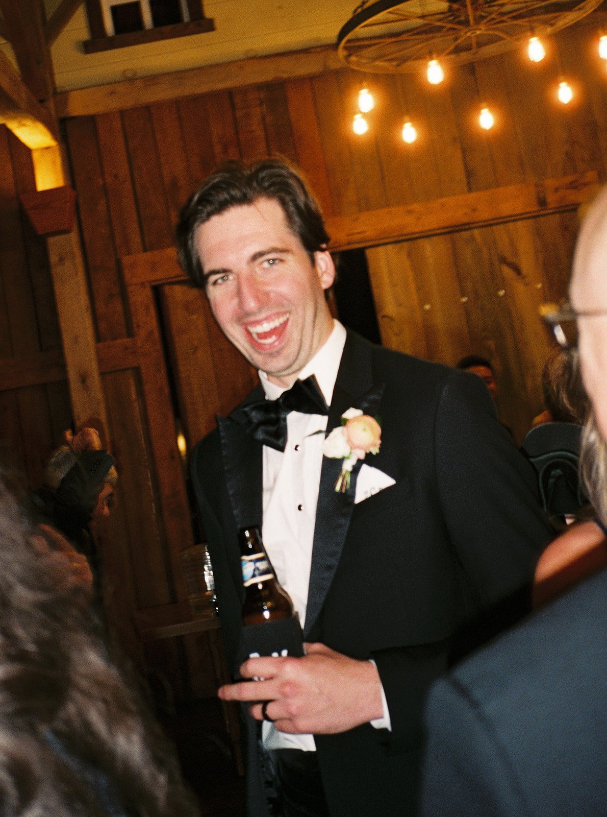 A man in a tuxedo with a bowtie and boutonniere, smiling and holding a beer bottle at a social gathering in a wood-paneled room with warm lighting and hanging bulbs.