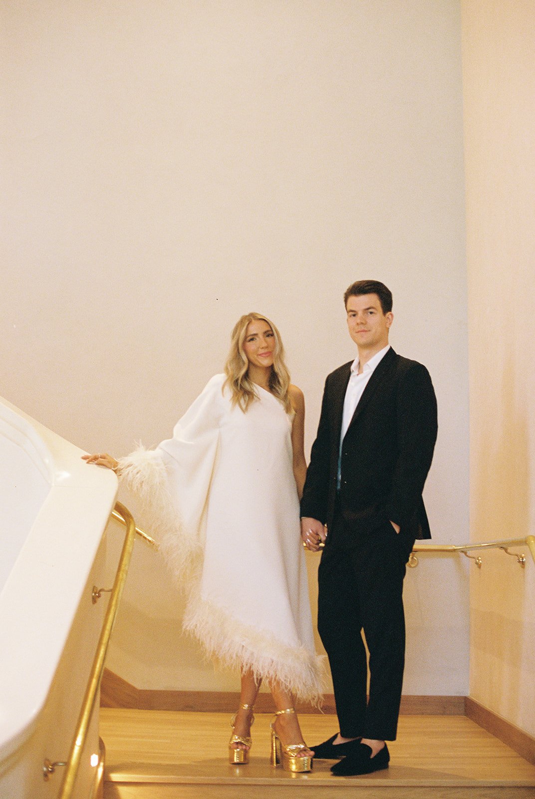 A woman in a white feather-trim dress and gold high heels stands with a man in a black suit and white shirt, holding hands, in an indoor setting with minimal decor and a staircase railings.
