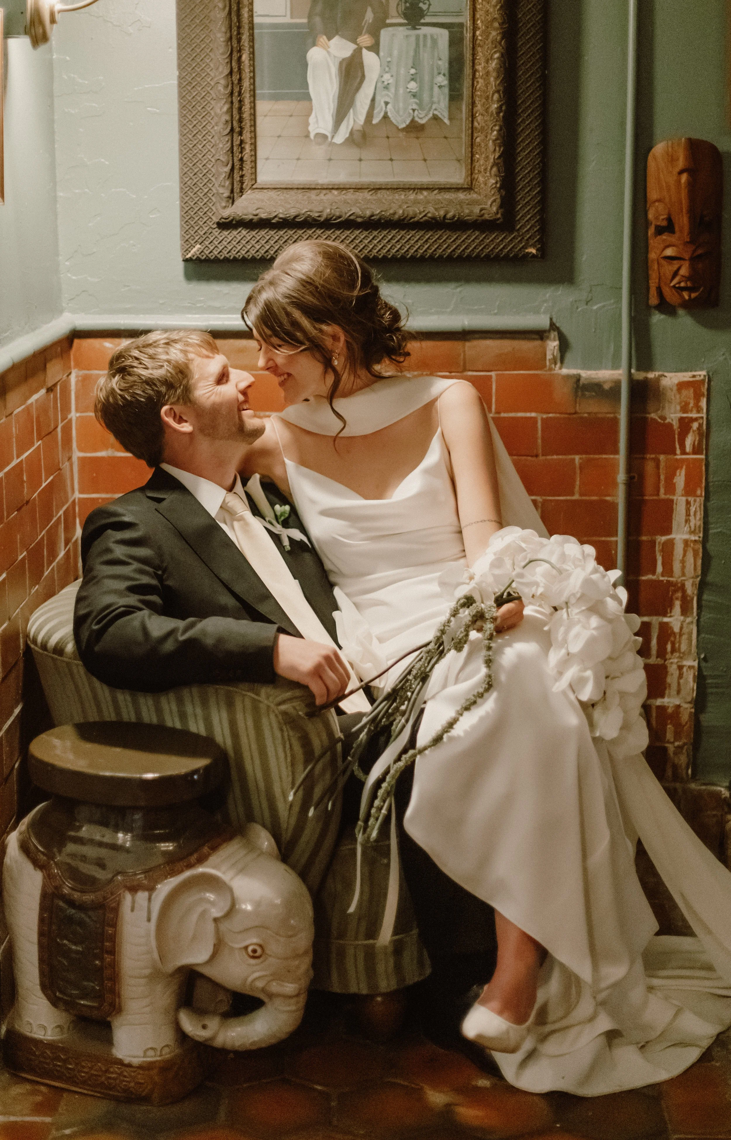 A couple on their wedding day, sitting on a vintage striped armchair; the bride is in a white wedding dress holding a bouquet, and the groom is in a black suit. They are smiling and leaning close to each other, in a cozy indoor setting with framed ar