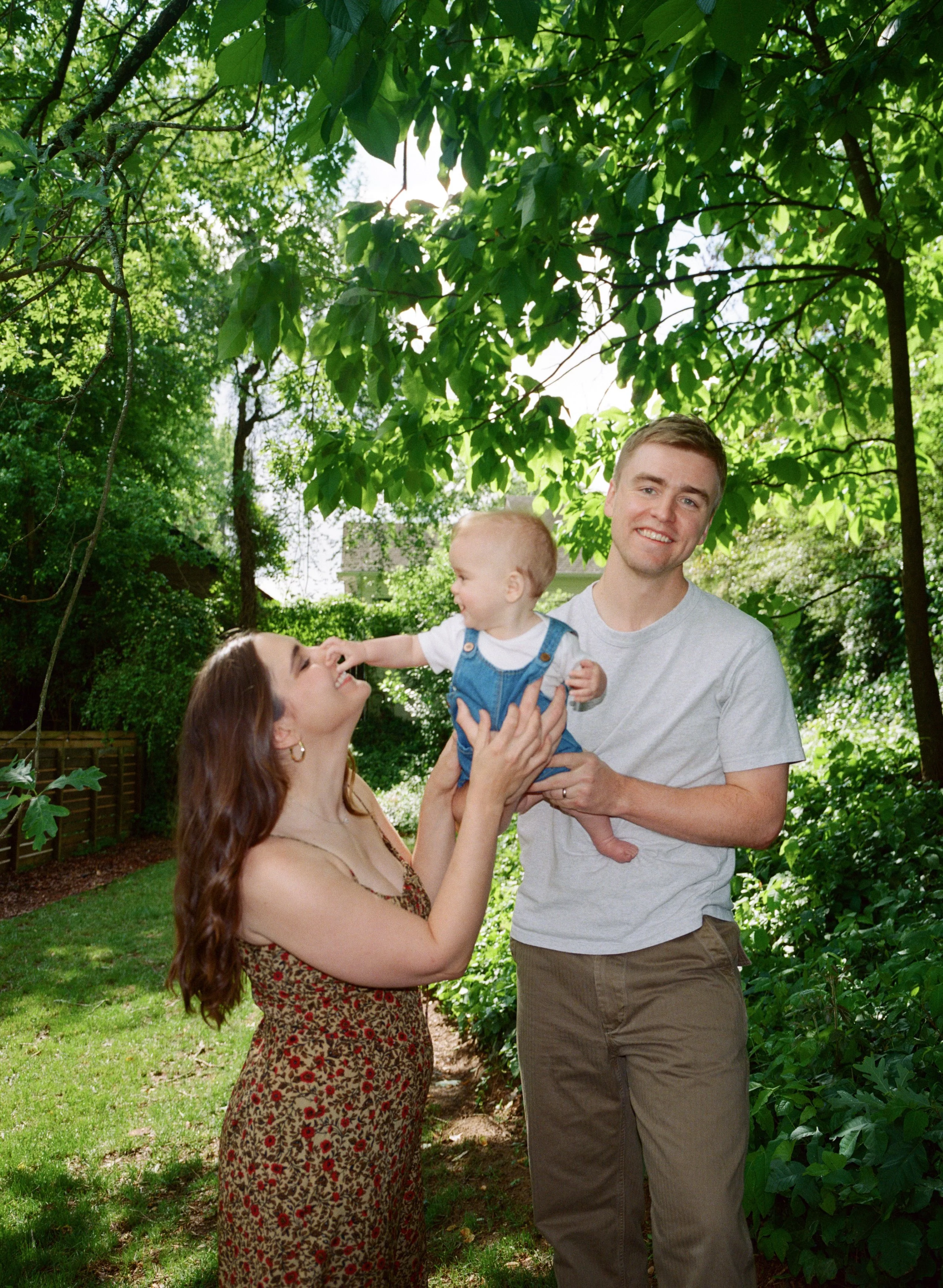 A smiling young family of three outdoors surrounded by green trees. The woman, with long brown hair, is wearing a floral dress and is lifting a baby boy, who is reaching towards her face. The man, with short hair, is standing next to them, holding th
