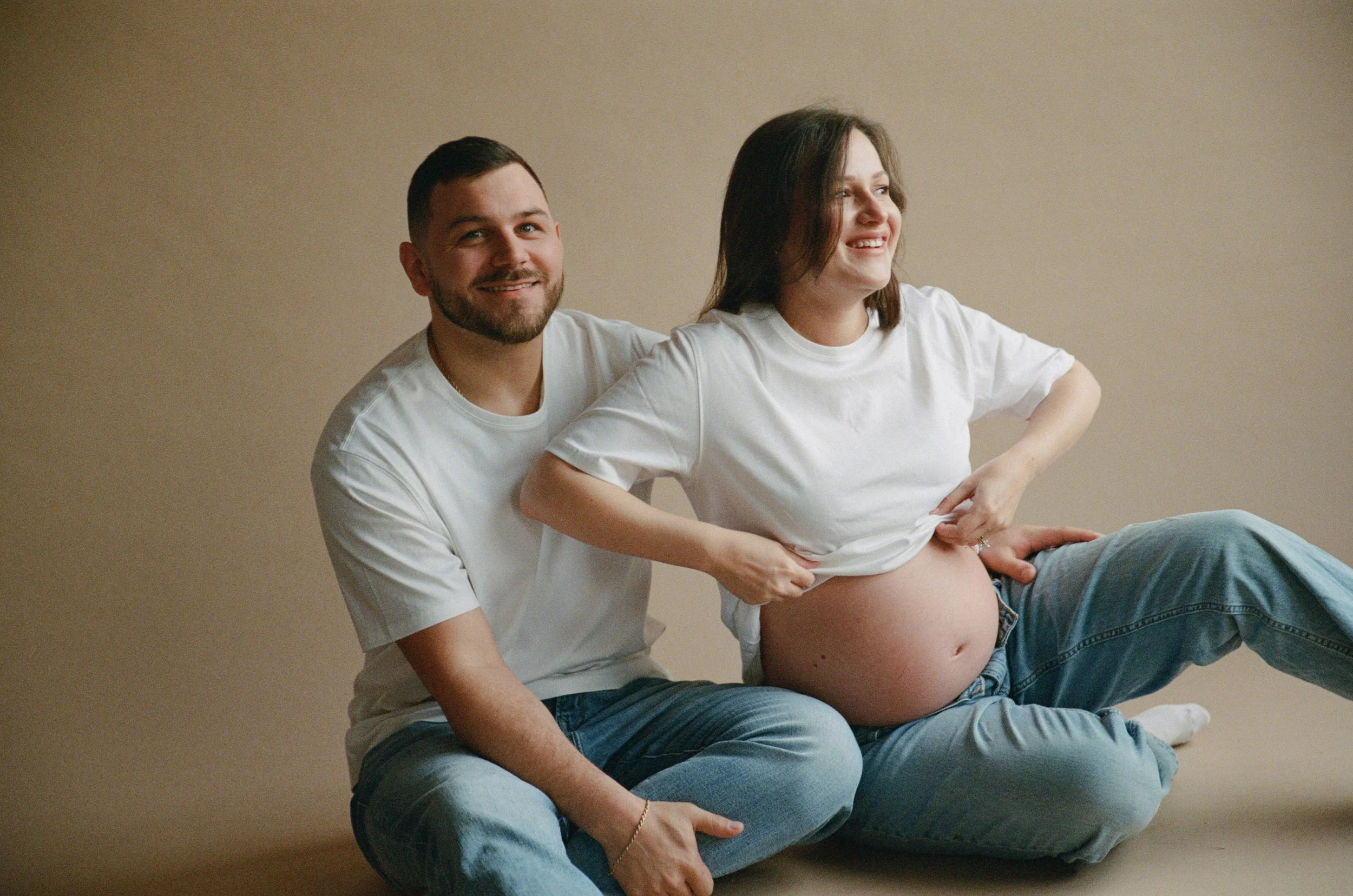 A pregnant woman with dark hair and a woman with short dark hair and a beard sitting on the floor against a beige background, smiling and looking away while lifting her shirt to show her pregnant belly.