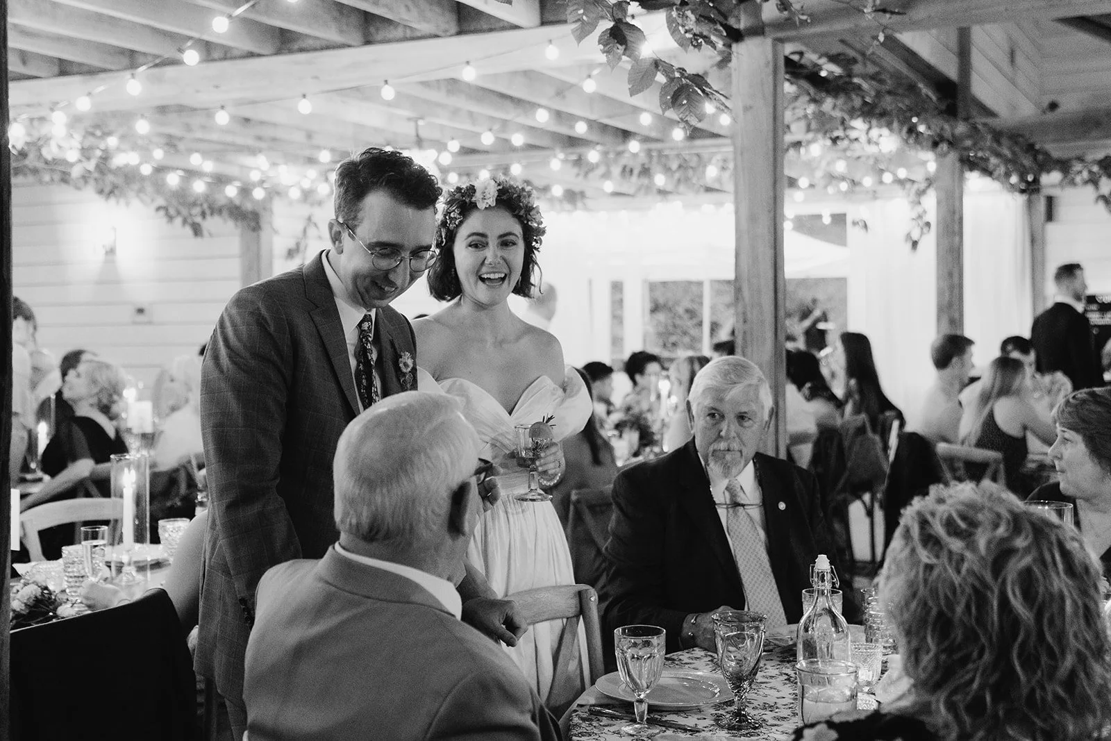 Black and white photograph of a wedding reception with a woman in a wedding dress and flower crown holding a drink, smiling with a man in a suit, and several guests seated at tables in a decorated venue.