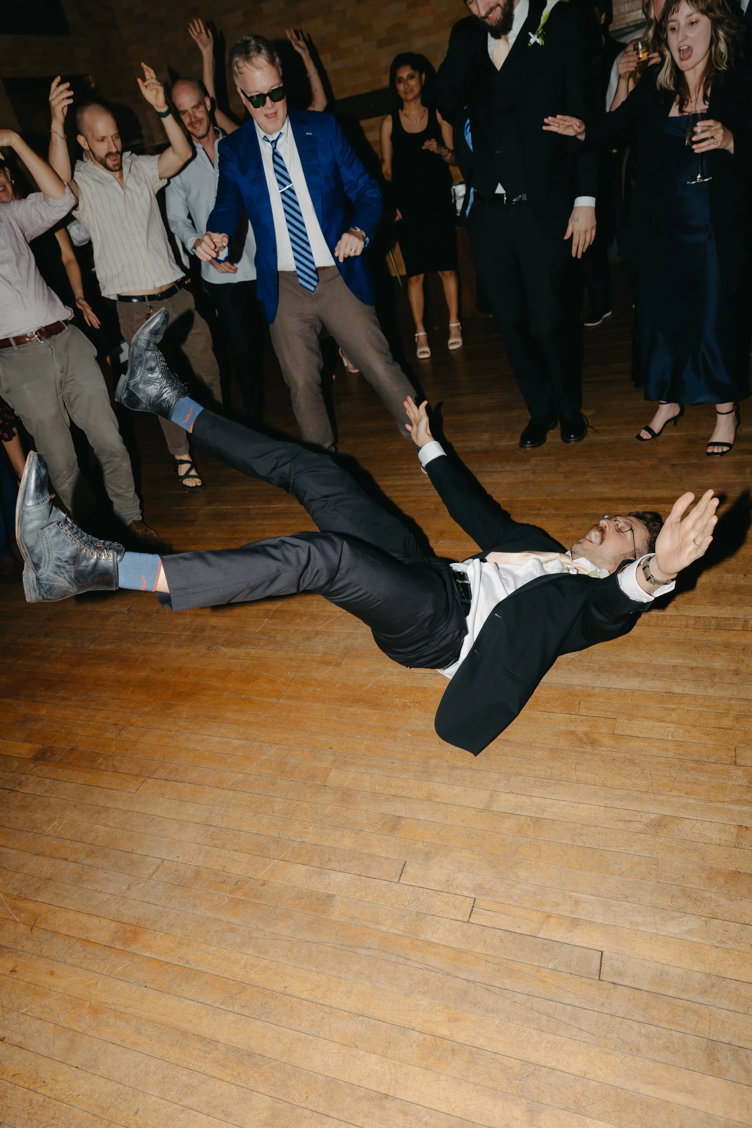 Man dressed in a tuxedo falling or being thrown on the dance floor during a celebration or party with people dancing and enjoying themselves.