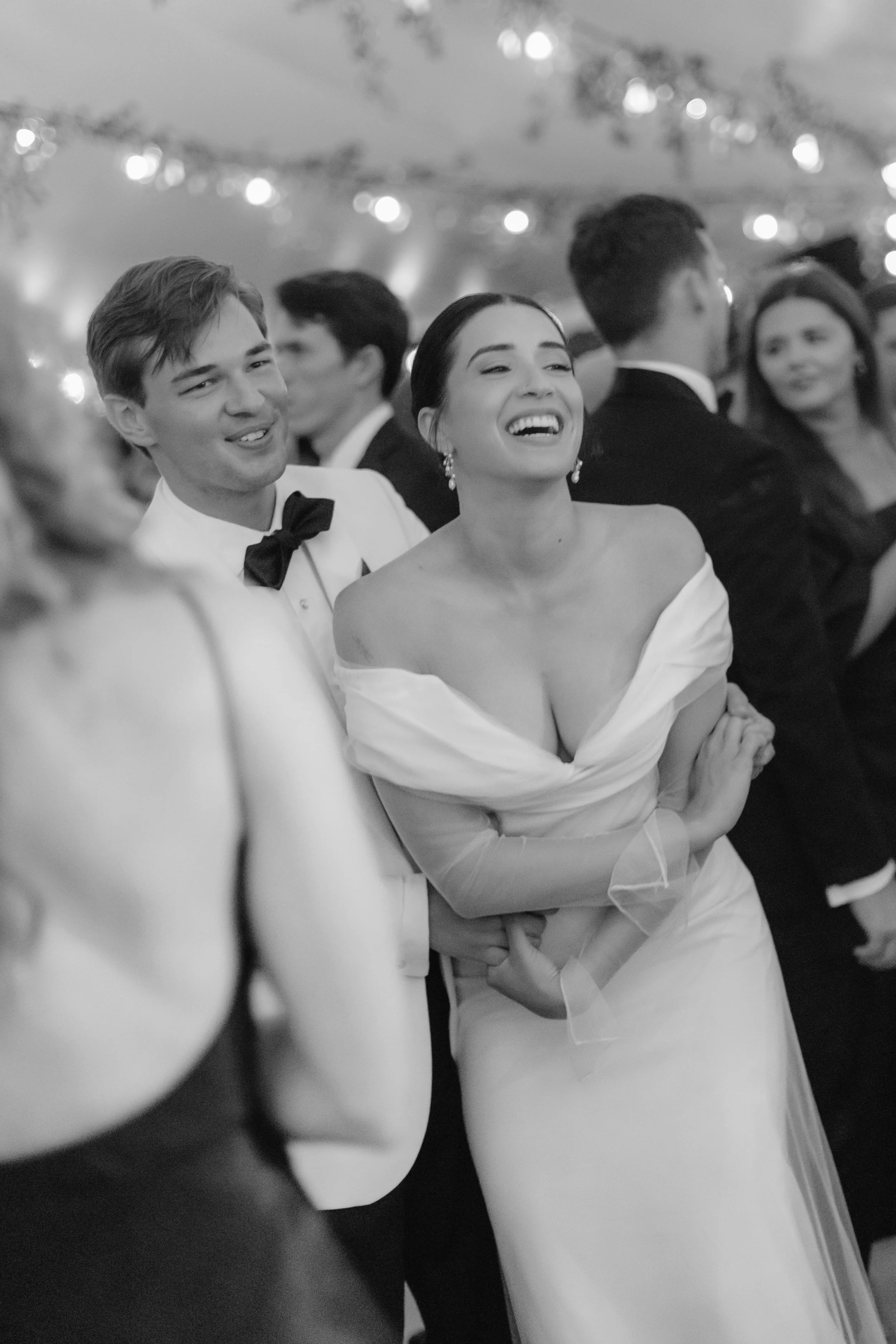 A group of people at a wedding reception, including a smiling woman in a wedding dress and a man in a tuxedo with a bow tie, dancing and enjoying the celebration.