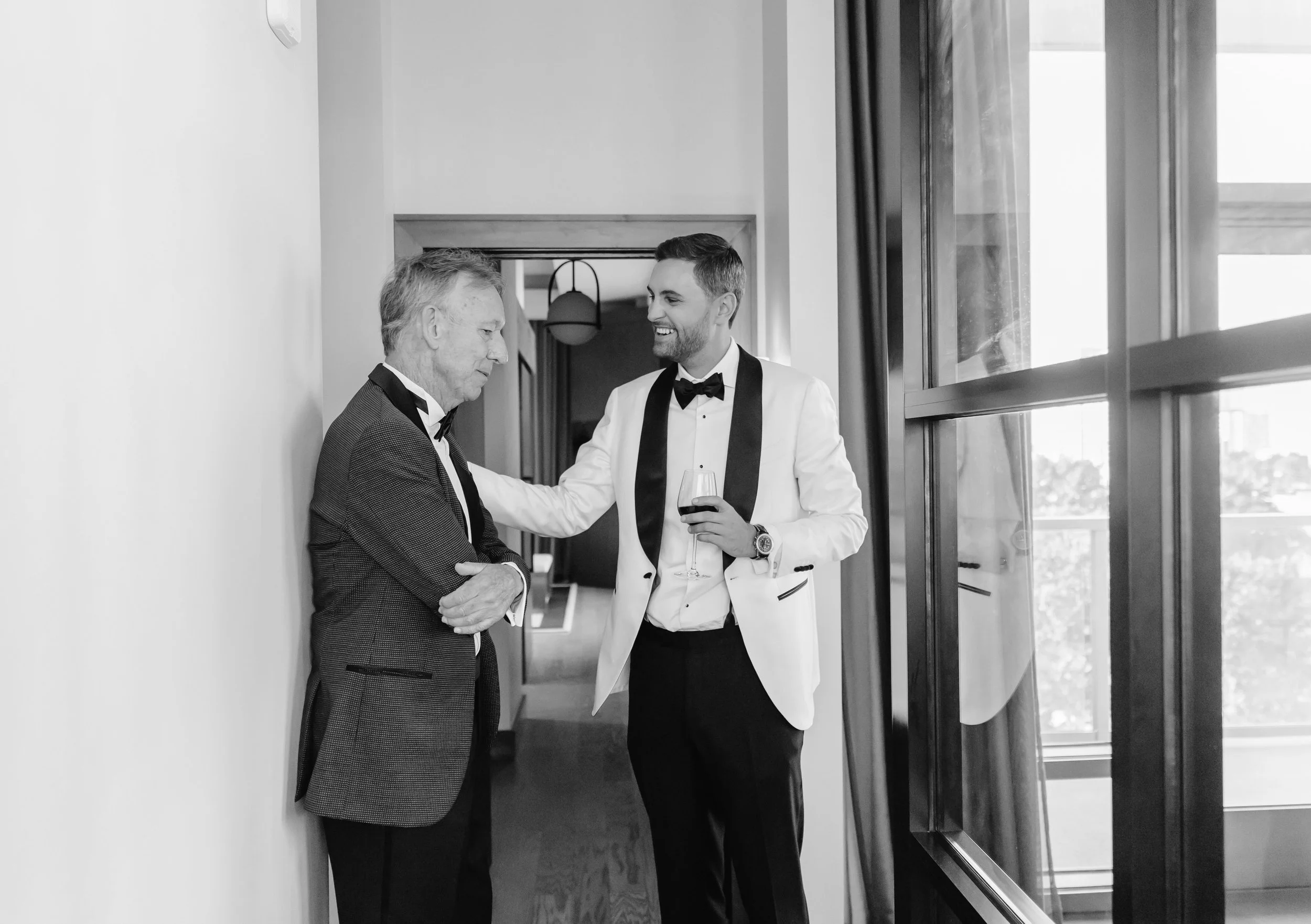 Two men in tuxedos having a conversation in a bright room with large windows, one with a glass of red wine.