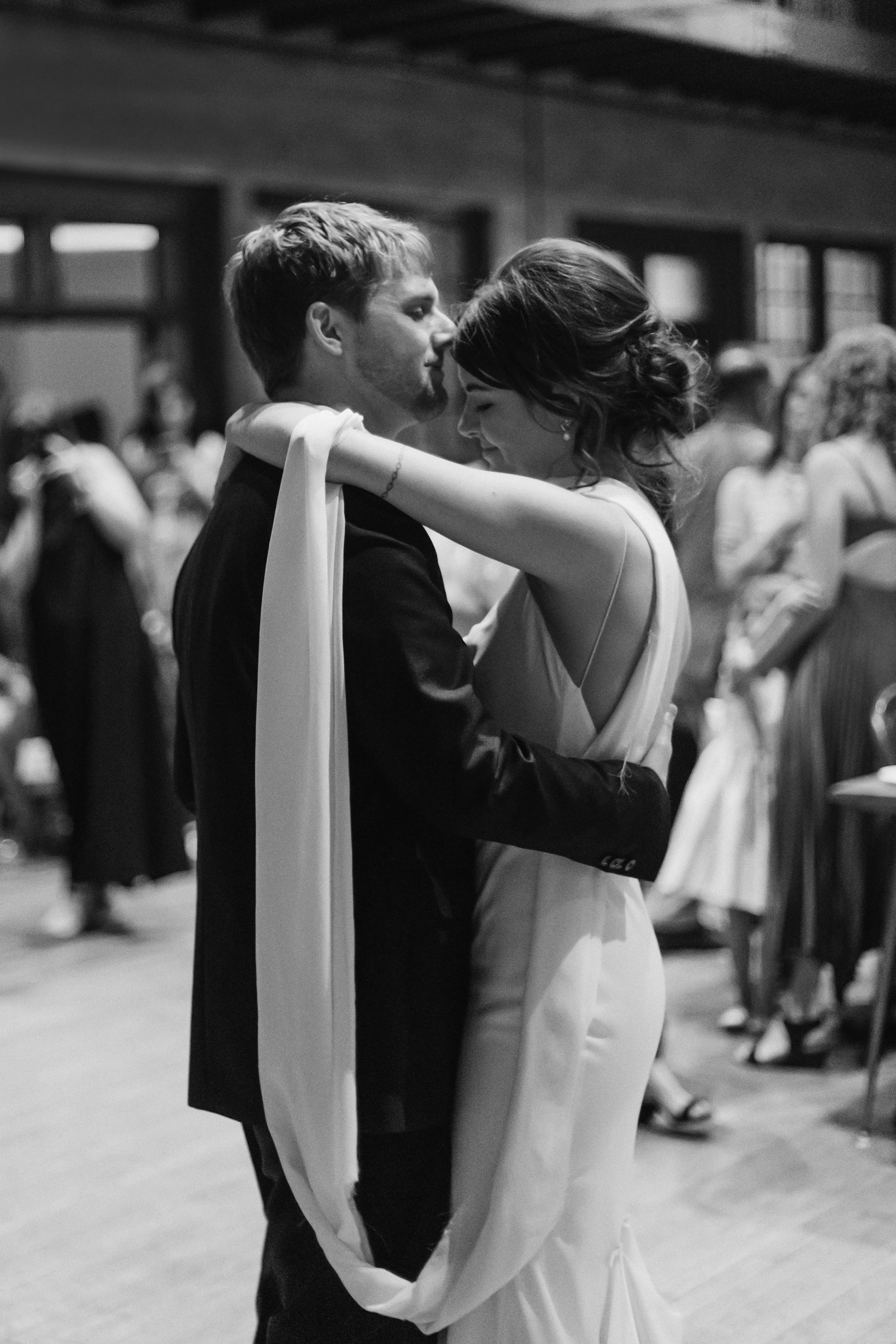 A black and white photo of a bride and groom dancing closely at their wedding reception, with other guests in the background.
