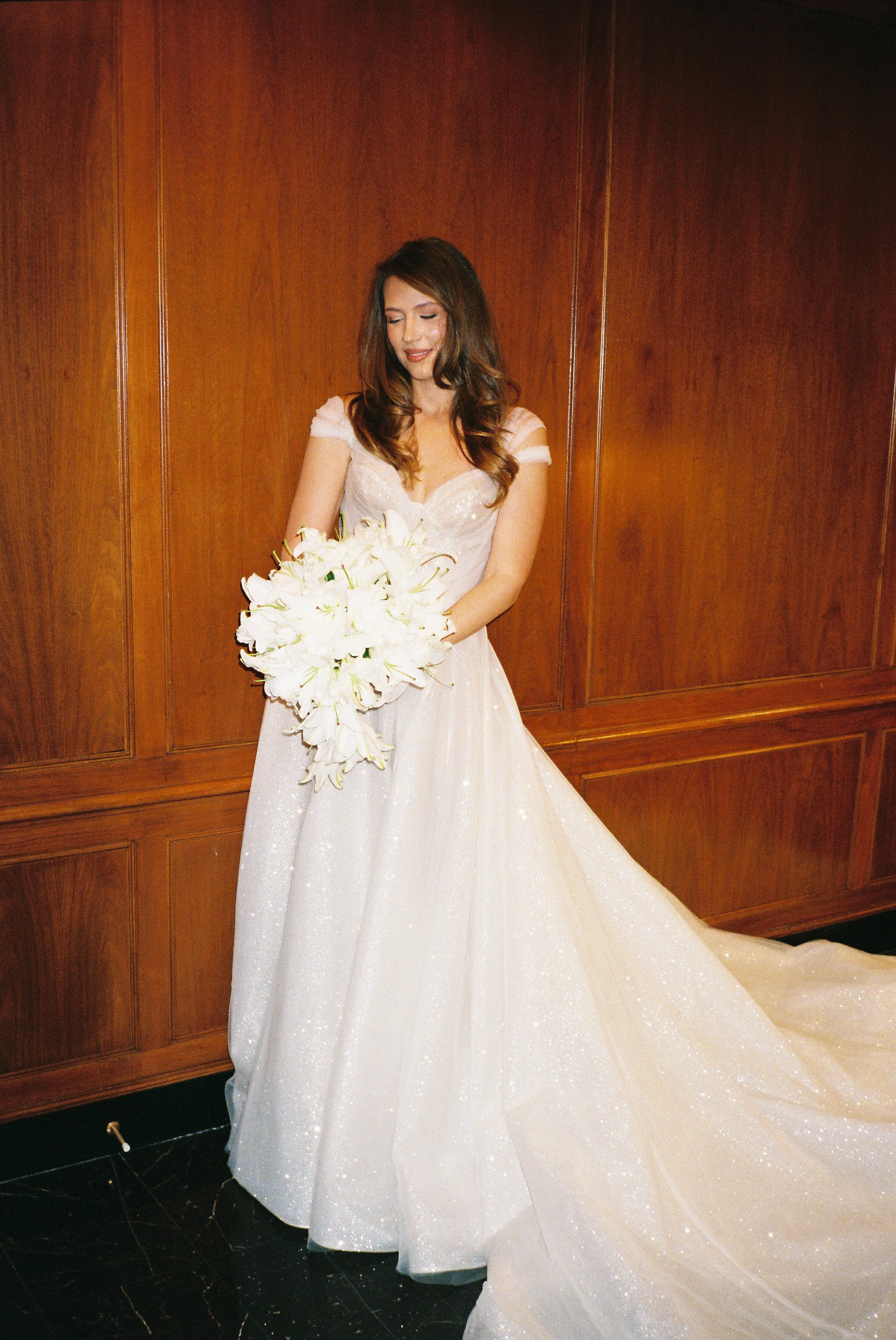 A woman in a wedding dress holding a bouquet of white flowers, standing against a wooden-paneled wall.