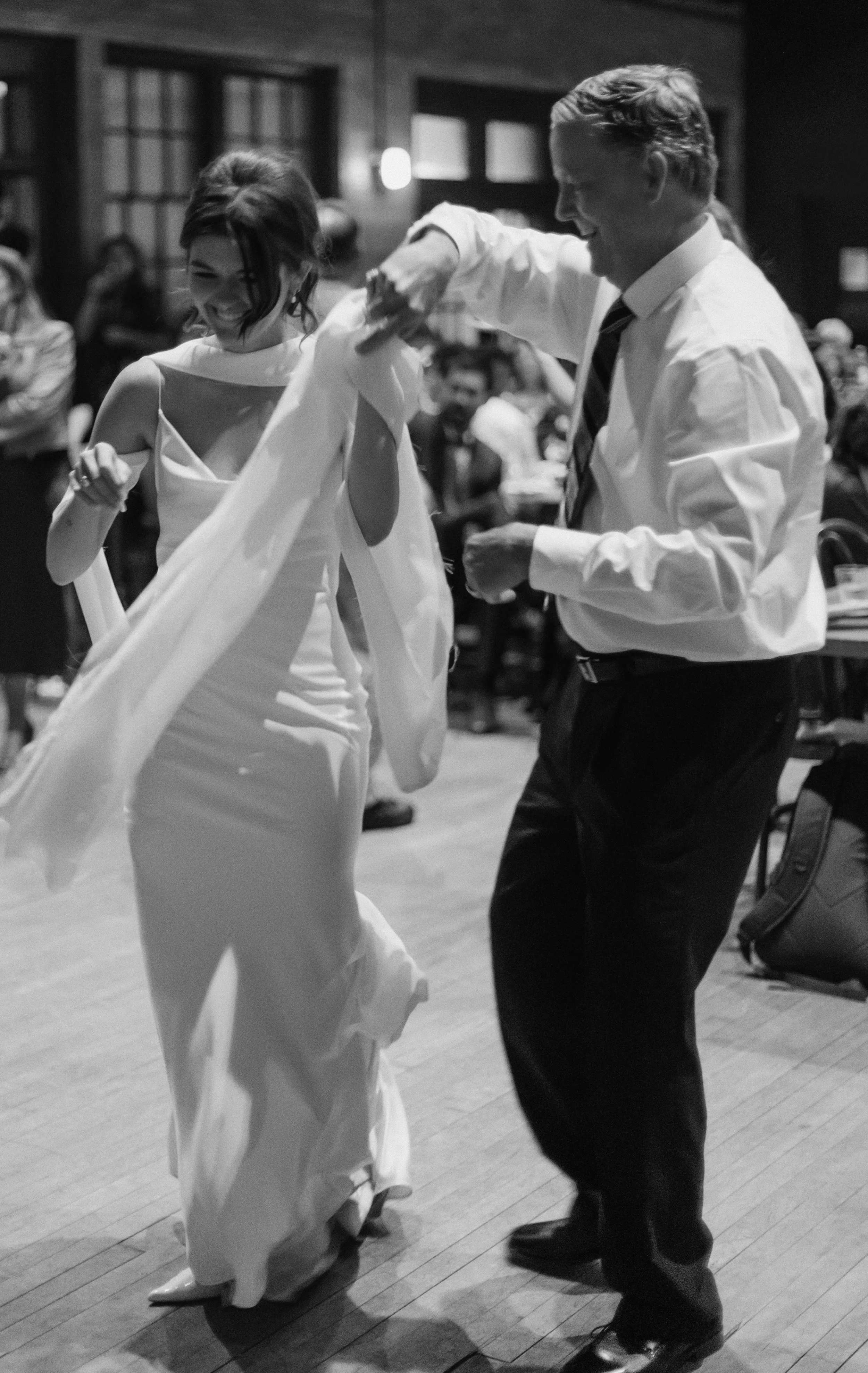 A black-and-white photo of a couple dancing at a wedding reception. The woman is wearing a white wedding dress and the man is dressed in a white shirt and dark pants. They are smiling and appear to be enjoying their dance.
