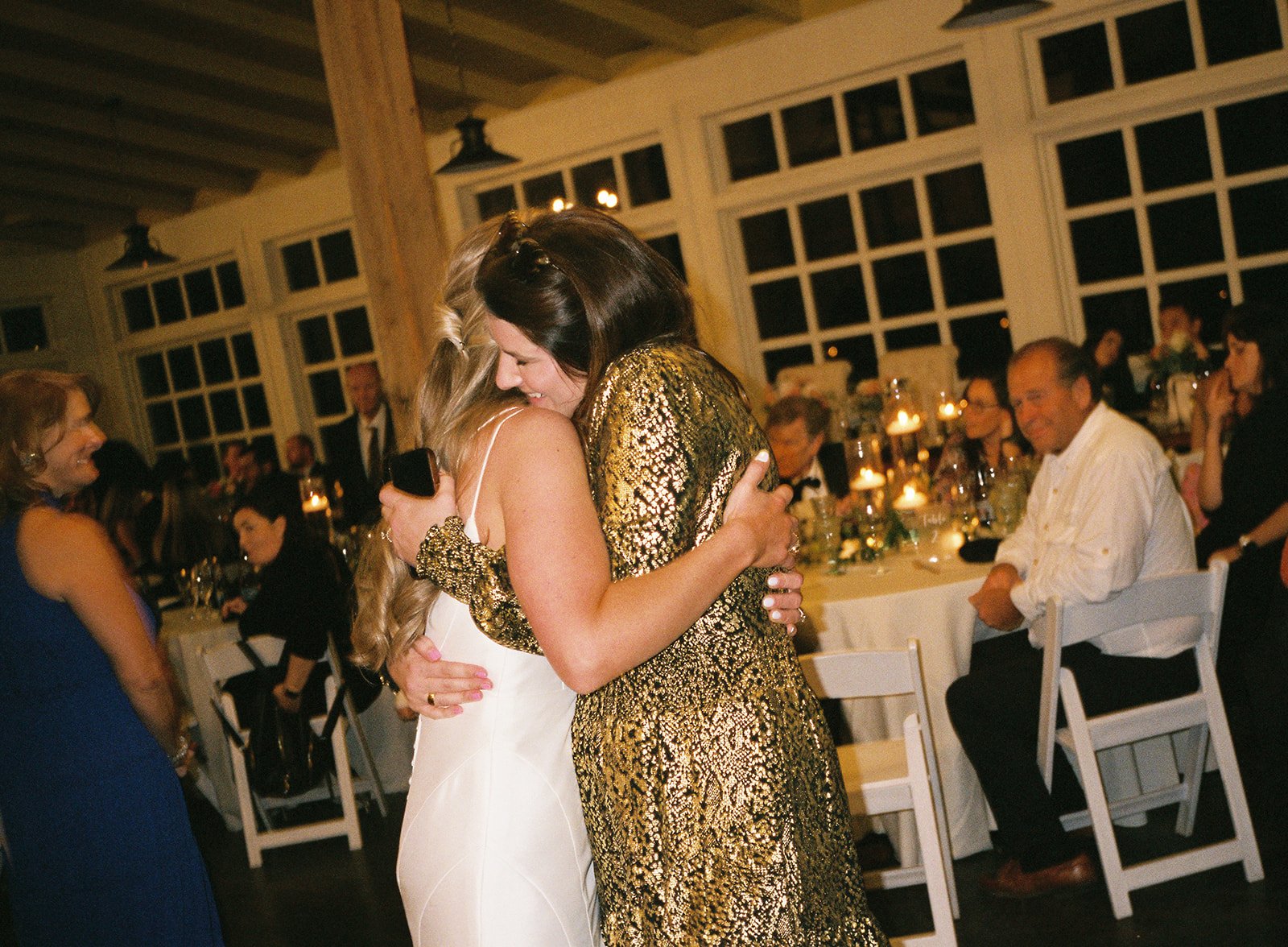 Two women hugging at a formal event, one in a white outfit and the other in a gold sequined dress, with guests seated at decorated tables in the background.