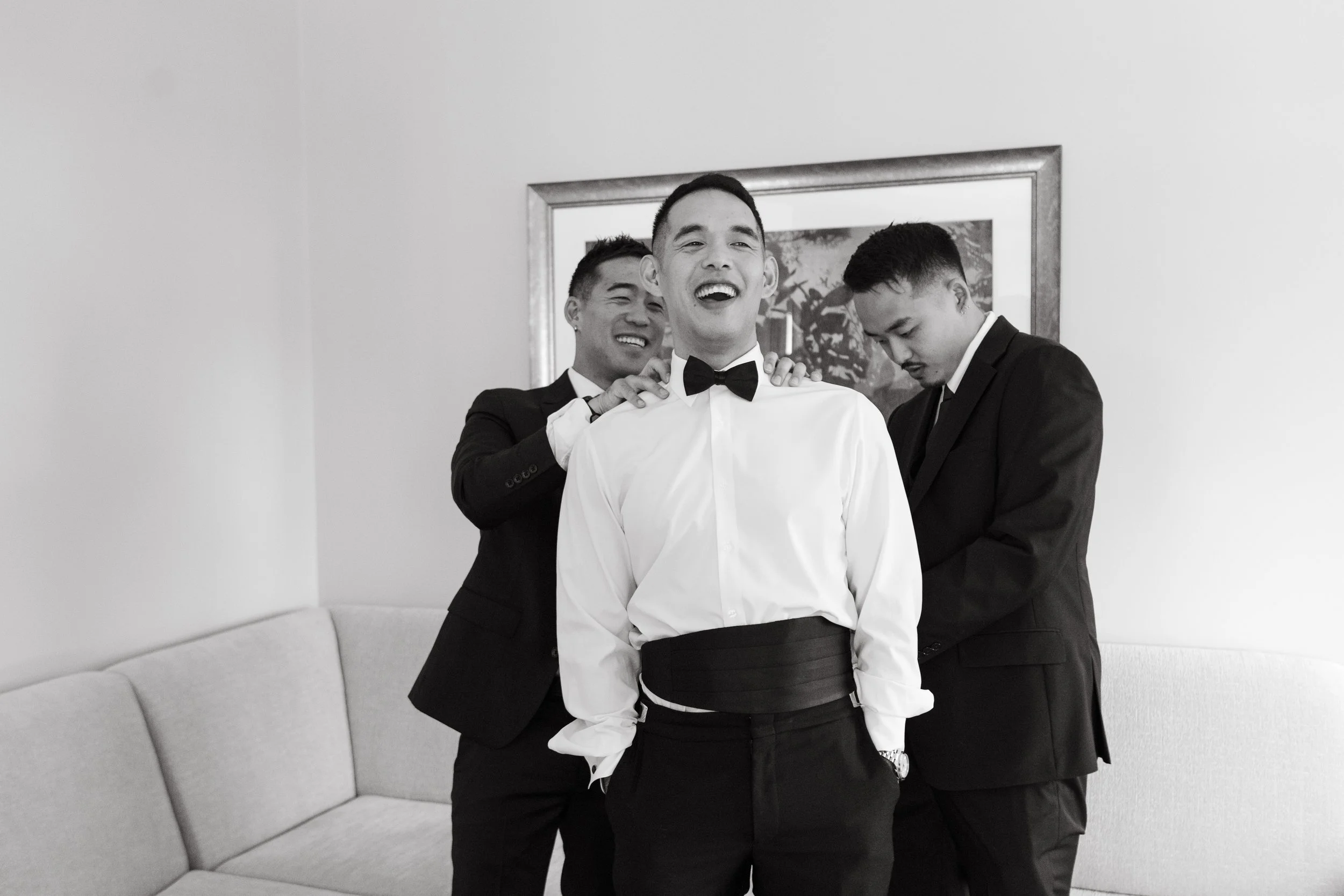 Three men in tuxedos, one with a bow tie, preparing for a formal event in a room with a framed picture on the wall and a couch, smiling and adjusting each other's attire.