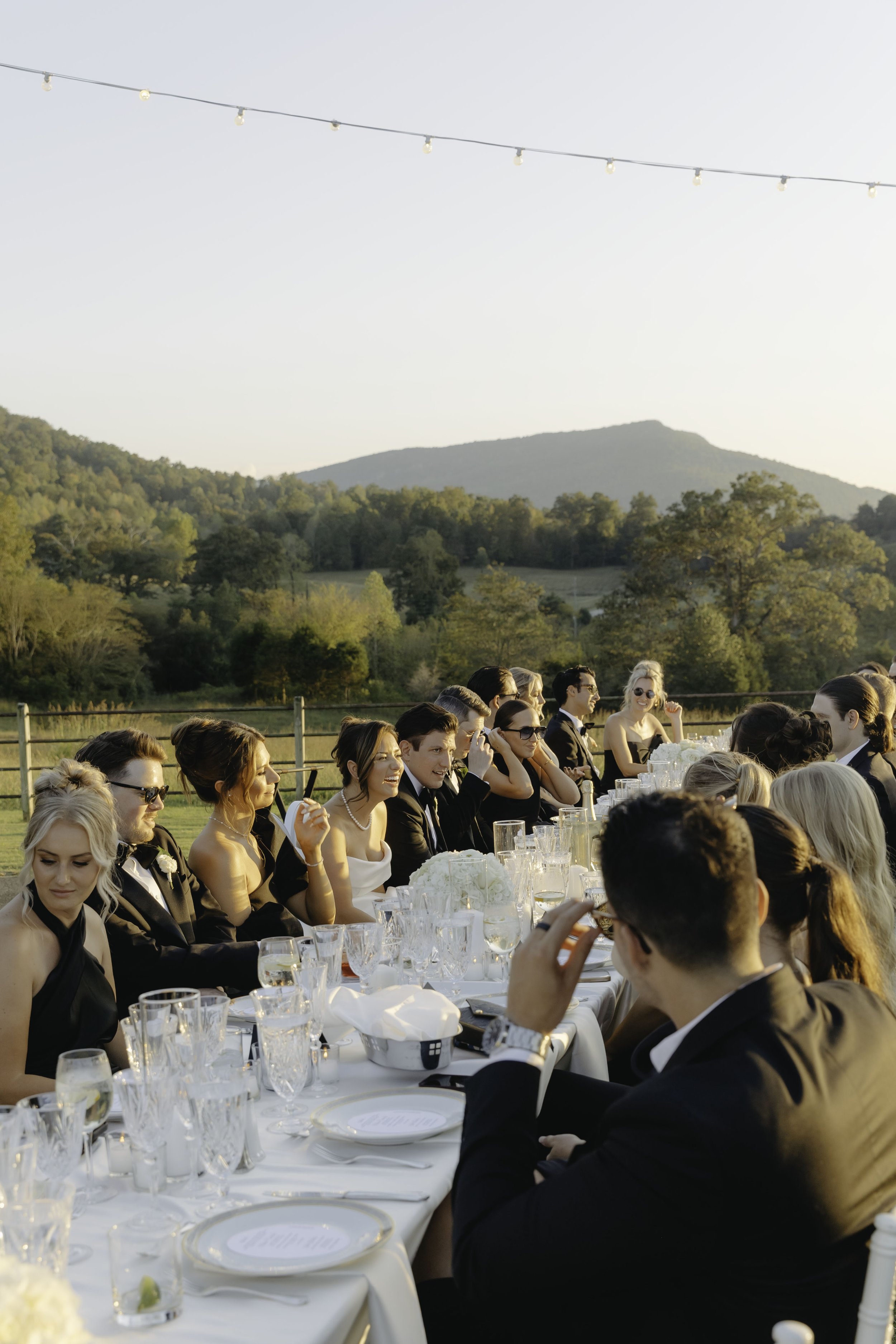 A large outdoor wedding reception with guests seated at a long table covered with a white tablecloth, set with glassware, plates, and floral centerpieces, with a backdrop of green trees, rolling hills, and a mountain under a clear sky, during late af
