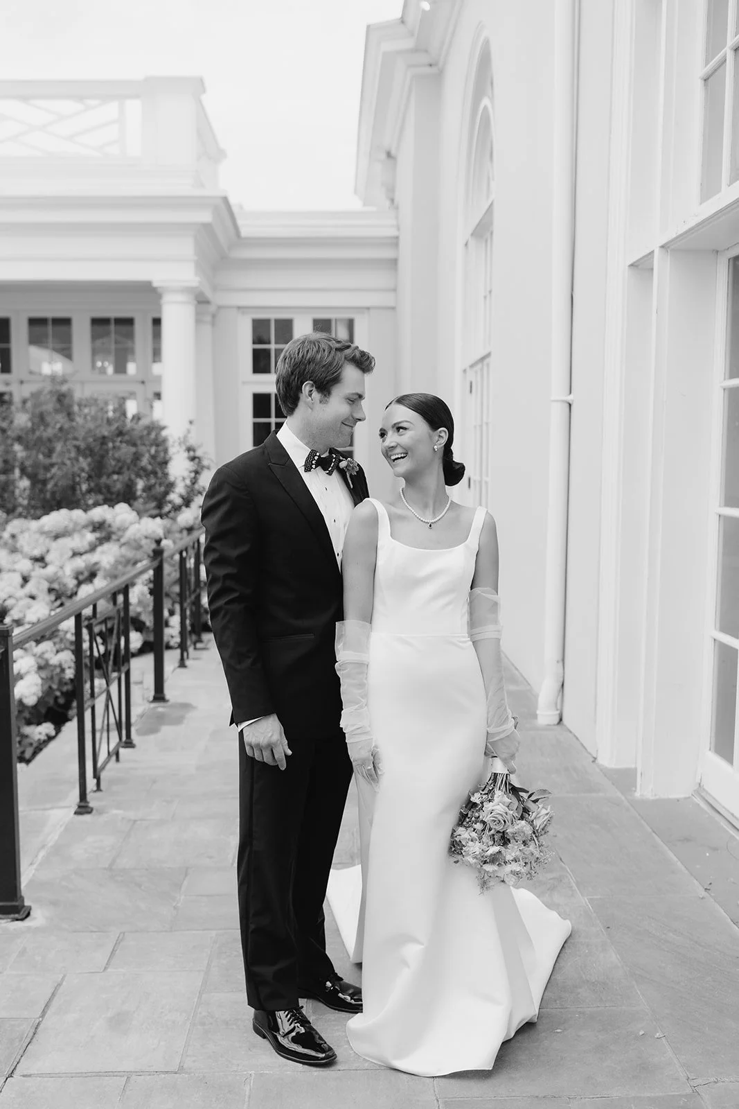 A bride and groom in wedding attire smiling at each other outdoors.