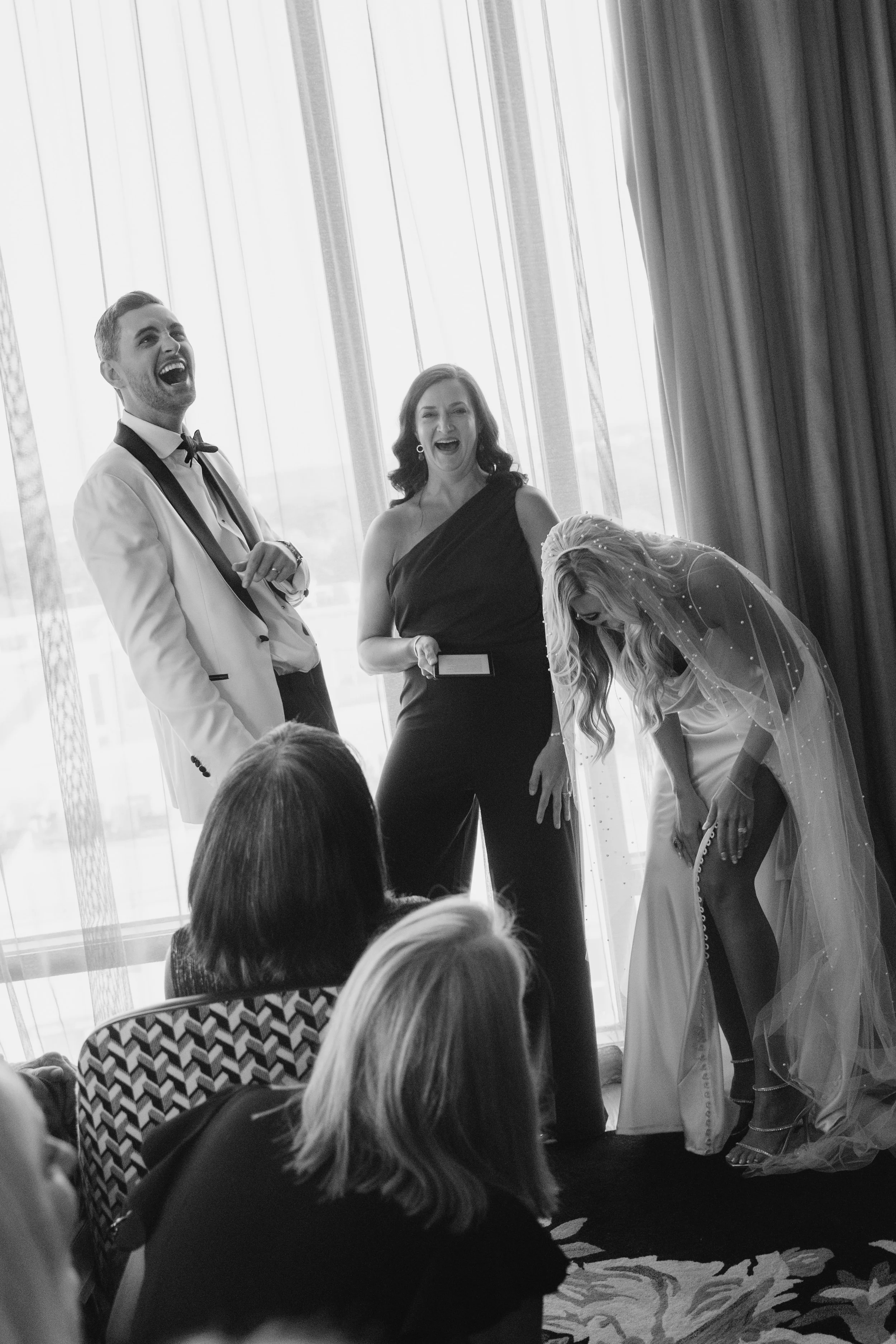 People at a wedding celebration, with a bride in a veil bending down, a woman holding a card, and a man and woman laughing.