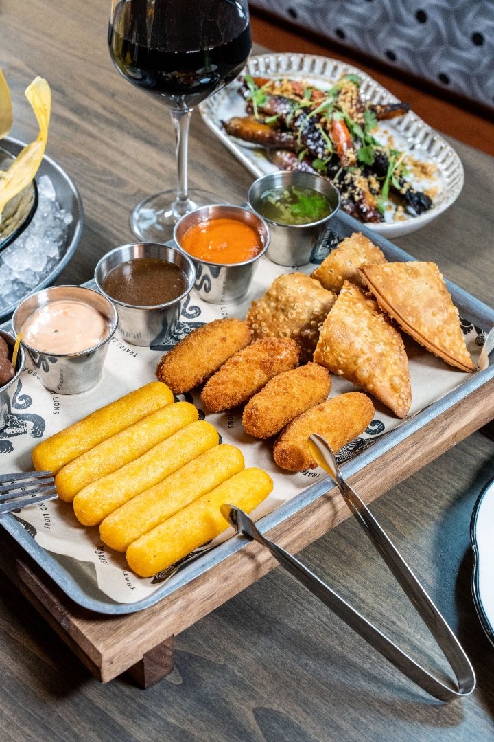 Tray with assorted appetizers, including empanadas, croquettes, and corn sticks, accompanied by dipping sauces; glass of red wine in background.