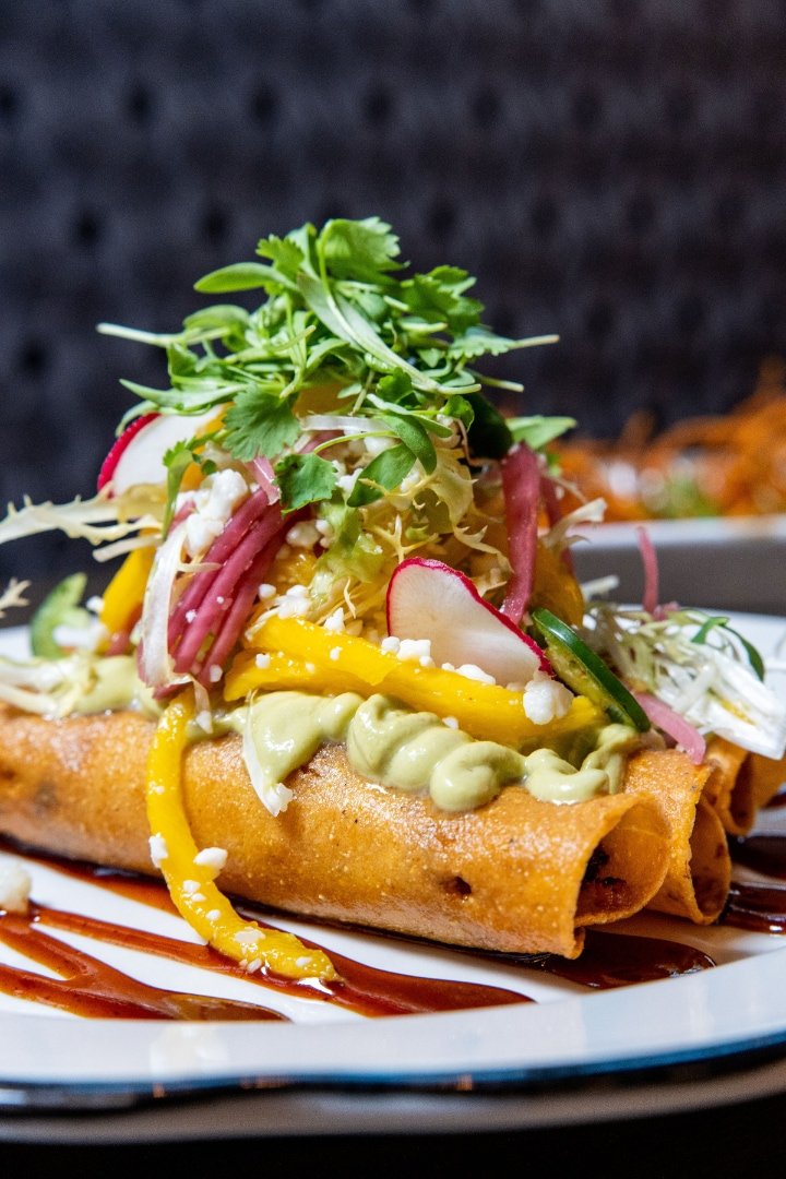 Crispy taquitos topped with avocado sauce, sliced radishes, pickled onions, fresh cilantro, shredded lettuce, and cheese on a plate with a drizzle of sauce.