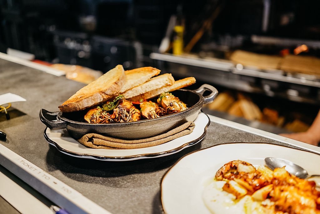 A dish of cooked shrimp with herbs and spices, served in a small cast iron pan with two slices of bread on top. Another plate of food is visible in the foreground on a kitchen countertop.