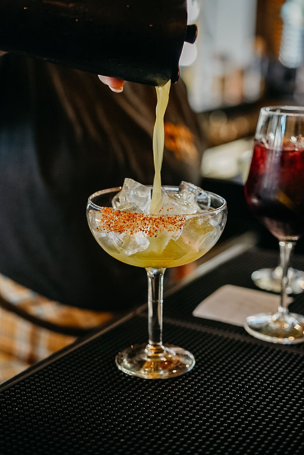 A bartender pouring a yellow cocktail into a glass with ice and a chili powder rim on a bar counter.