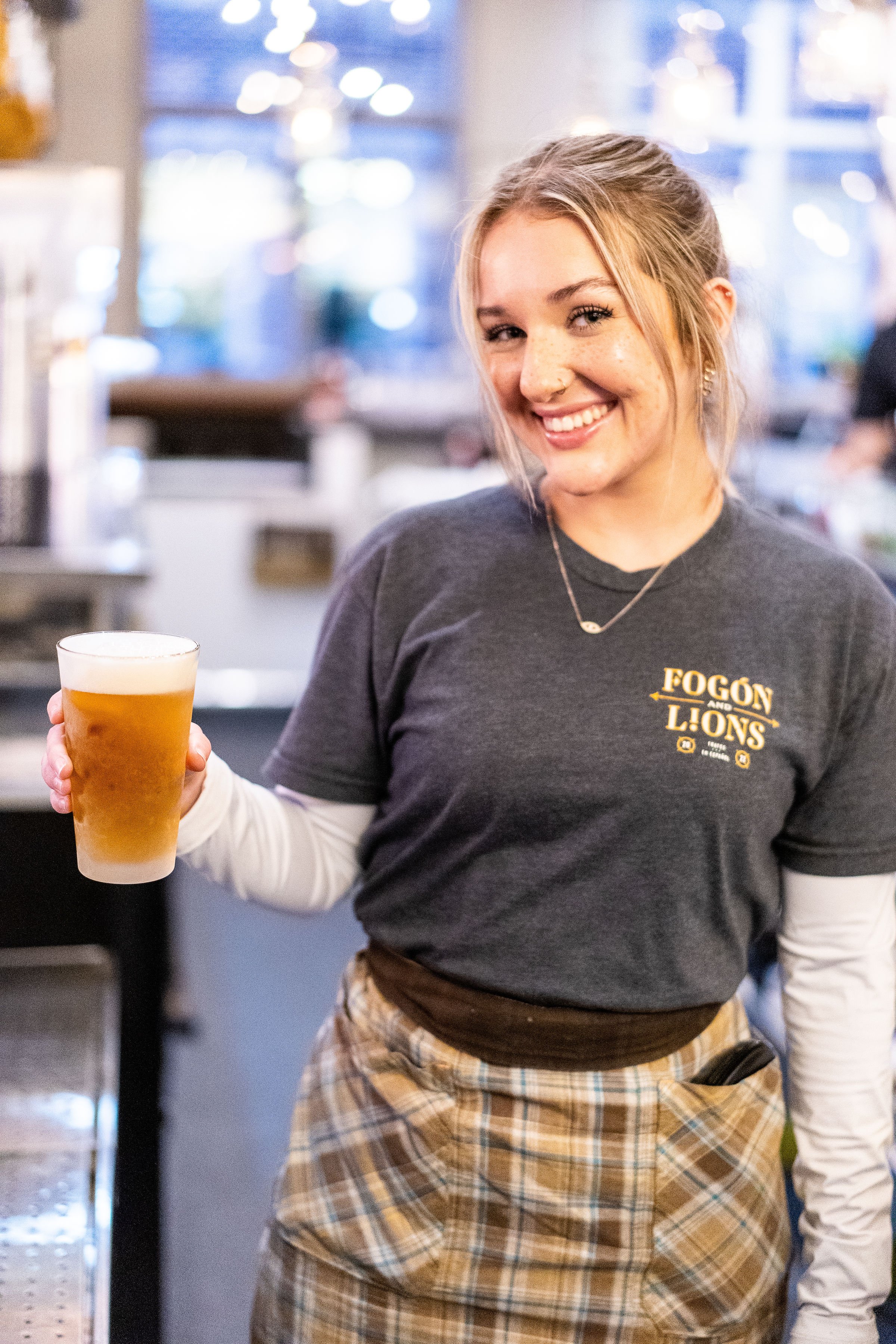 A smiling woman holding a glass of beer at a bar or restaurant, wearing a "Fogón and Lions" shirt and a plaid apron, with a blurry background.