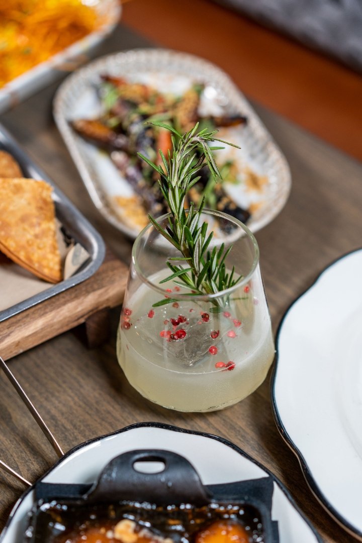 Close-up of a cocktail garnished with rosemary and pink peppercorns, surrounded by plates of food on a wooden table.