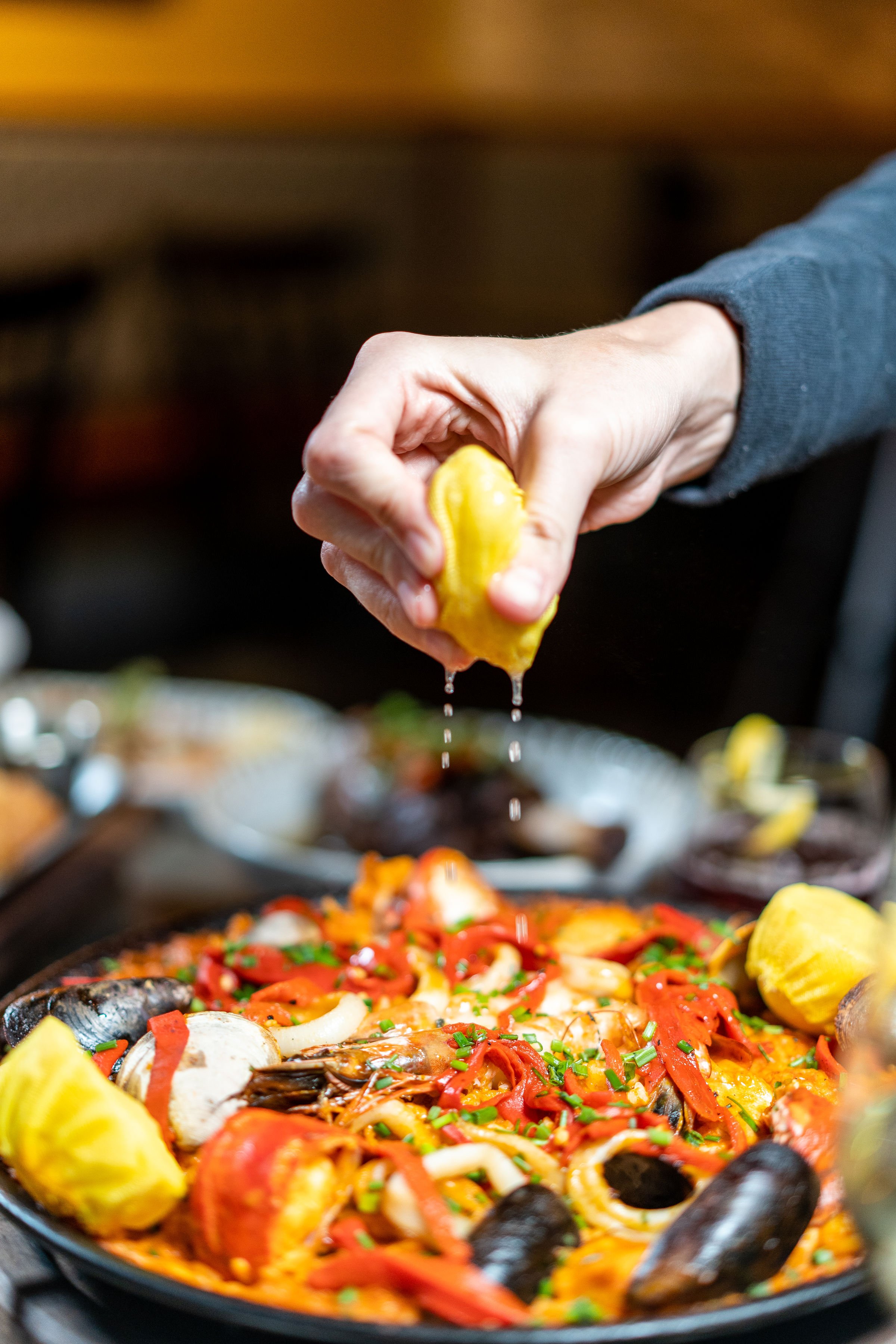 Person squeezing lemon over a mixed seafood paella with shrimp, mussels, and red peppers in a pan.