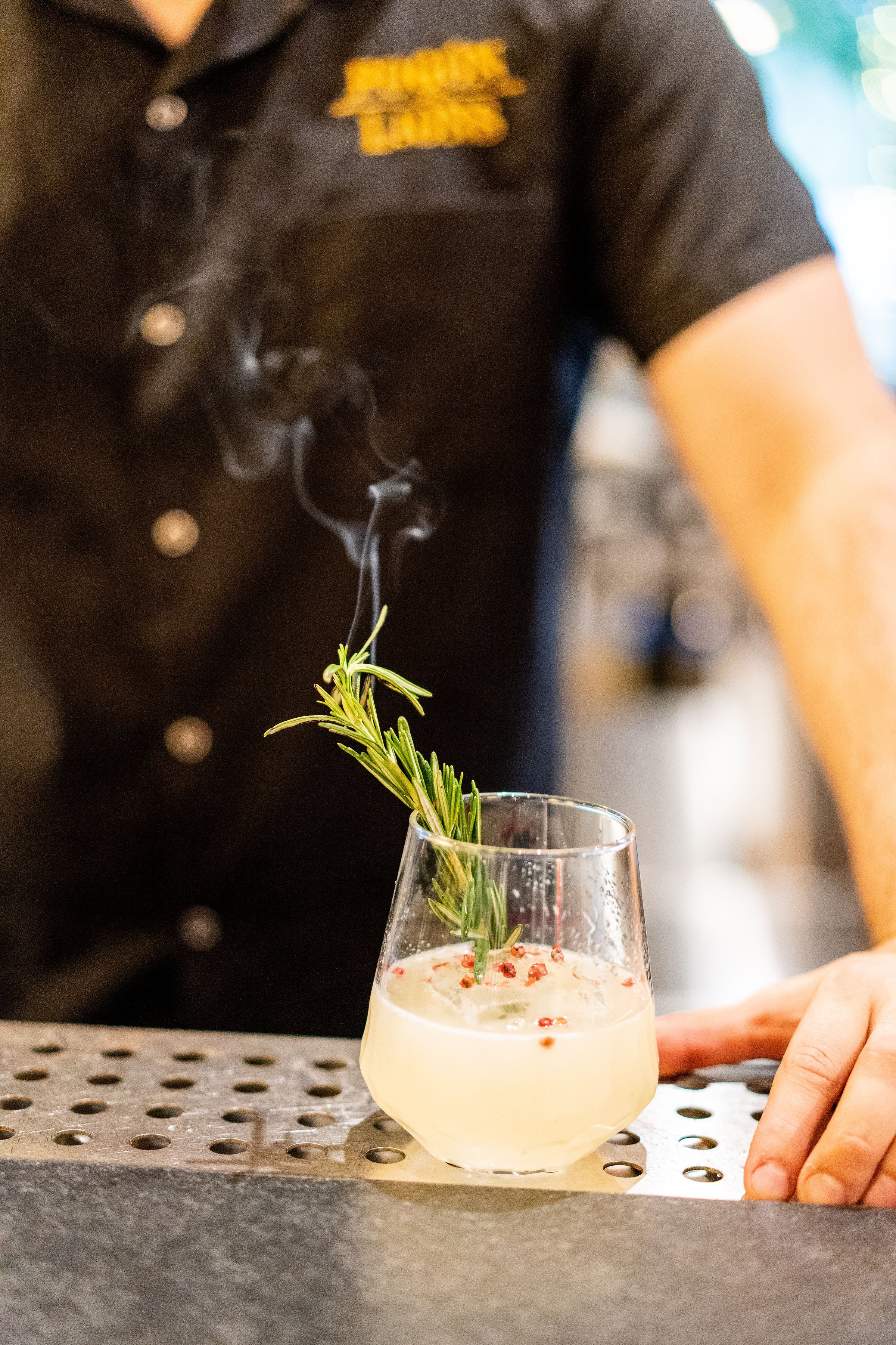 Smoky cocktail garnished with rosemary, served in a glass with ice and red peppercorns, held by a person in a black shirt.