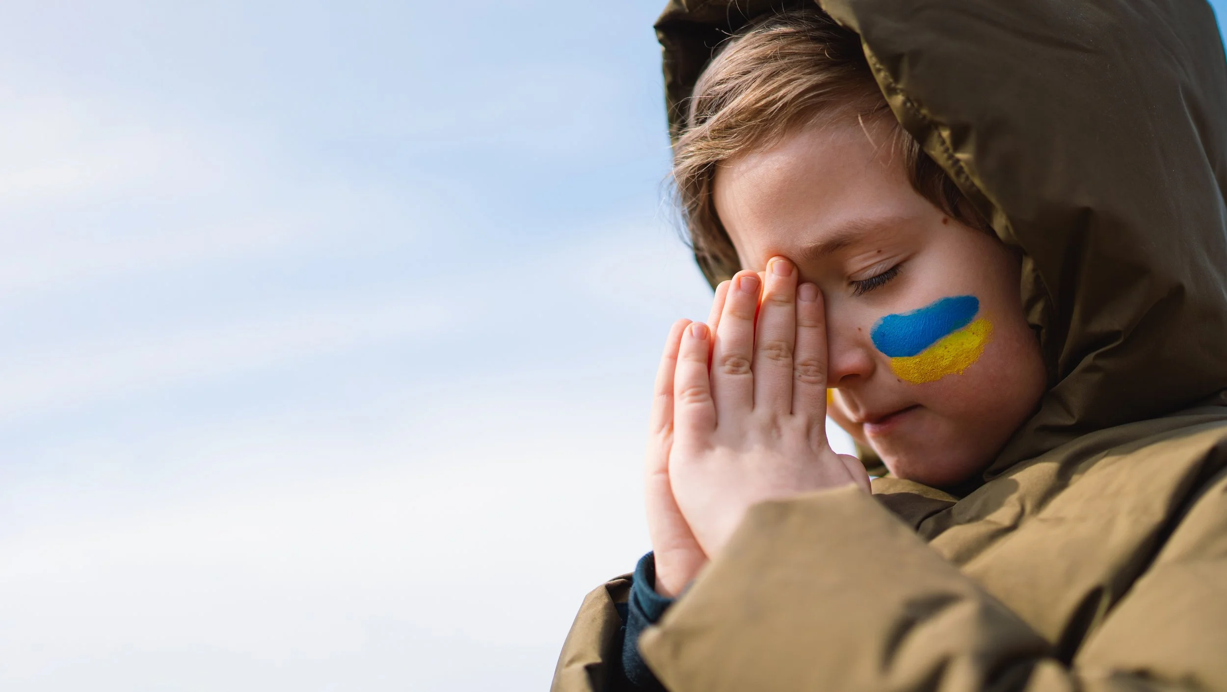 small child with blue and yellow stripes painted on their cheek, holding their hands up to their face in prayer hands