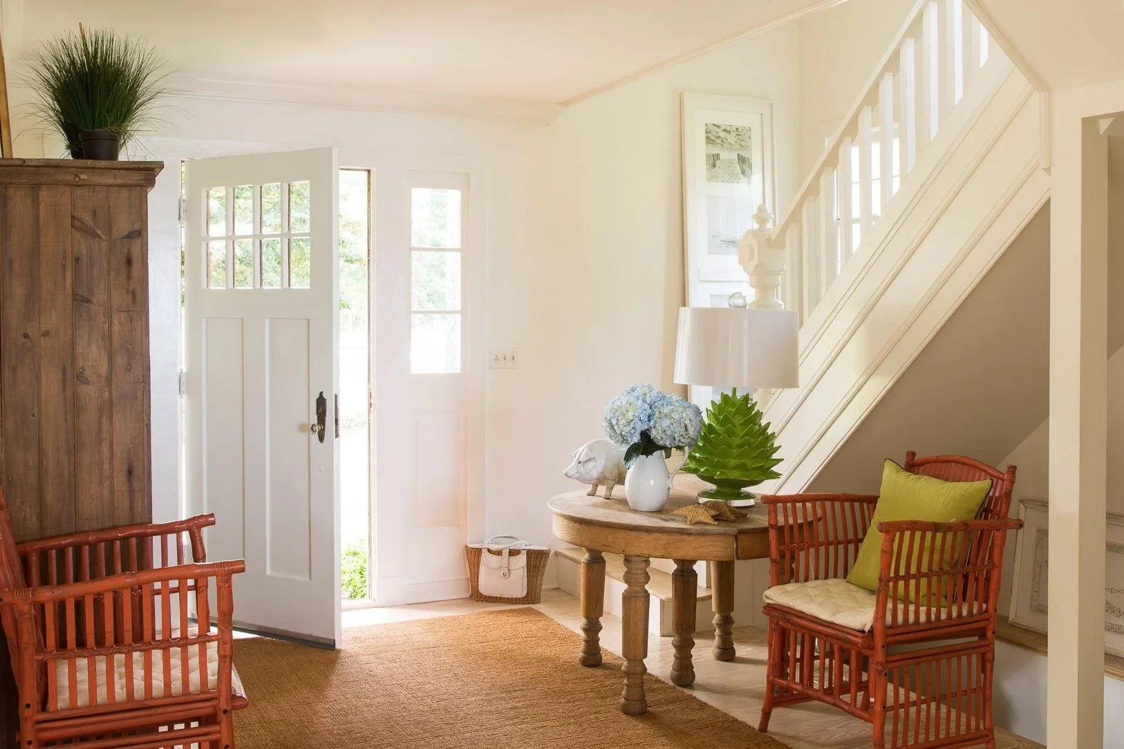 Entryway of a house with a white front door, a side staircase, a round wooden table decorated with a lamp, hydrangea flowers, a white pig figurine, a green leaf sculpture, and two rattan chairs with cushions.