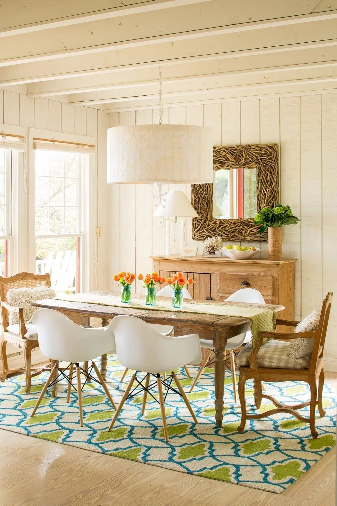 A bright dining room with white wooden walls and ceiling, a rustic wooden table with white and wooden chairs, colorful tulip centerpieces, a blue and green patterned rug, and a light wooden sideboard decorated with a mirror, a potted plant, and a bowl of green apples.