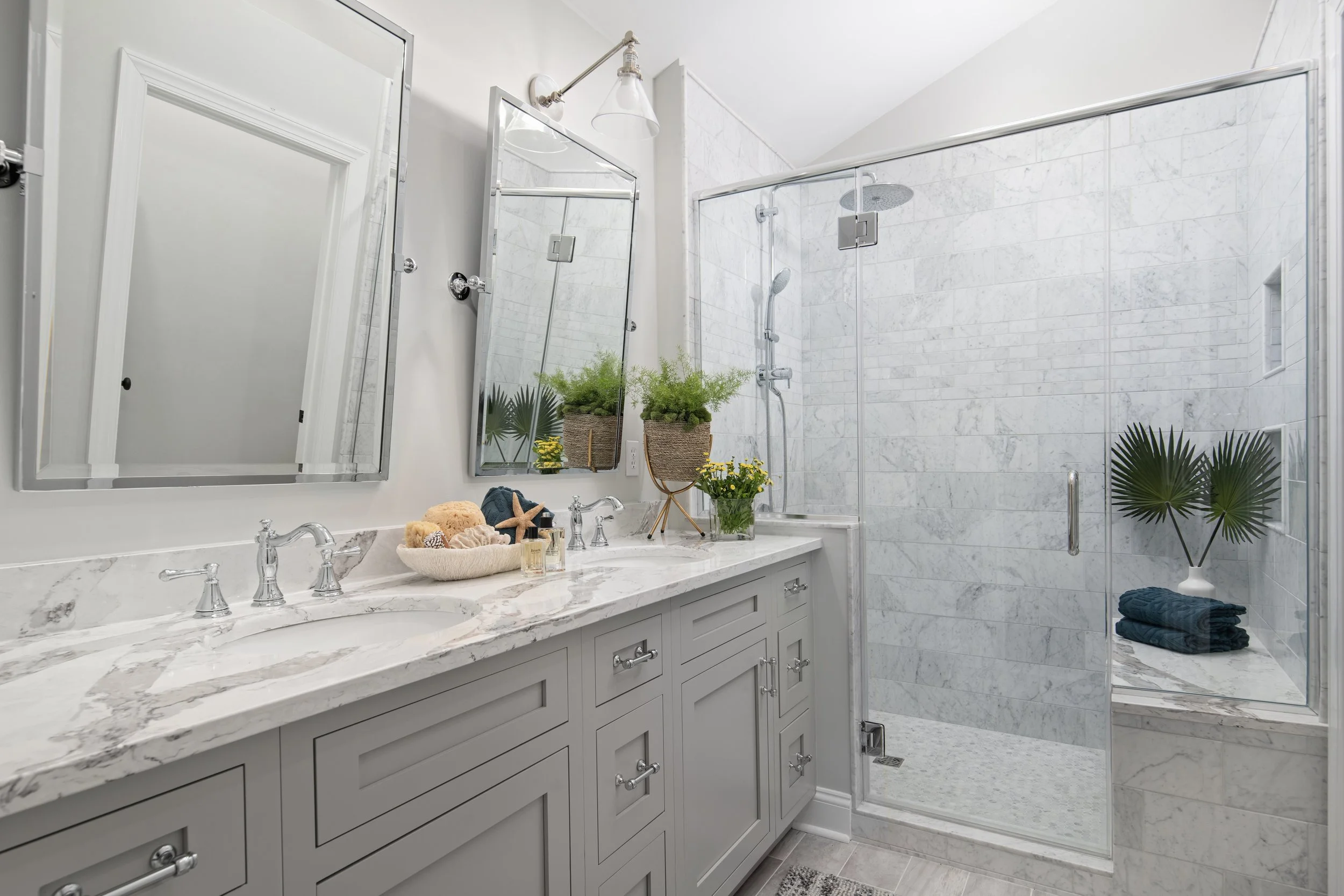 Modern bathroom with a white marble vanity, dual mirrors, and a large glass shower with marble walls. There are potted plants and folded towels for decoration.