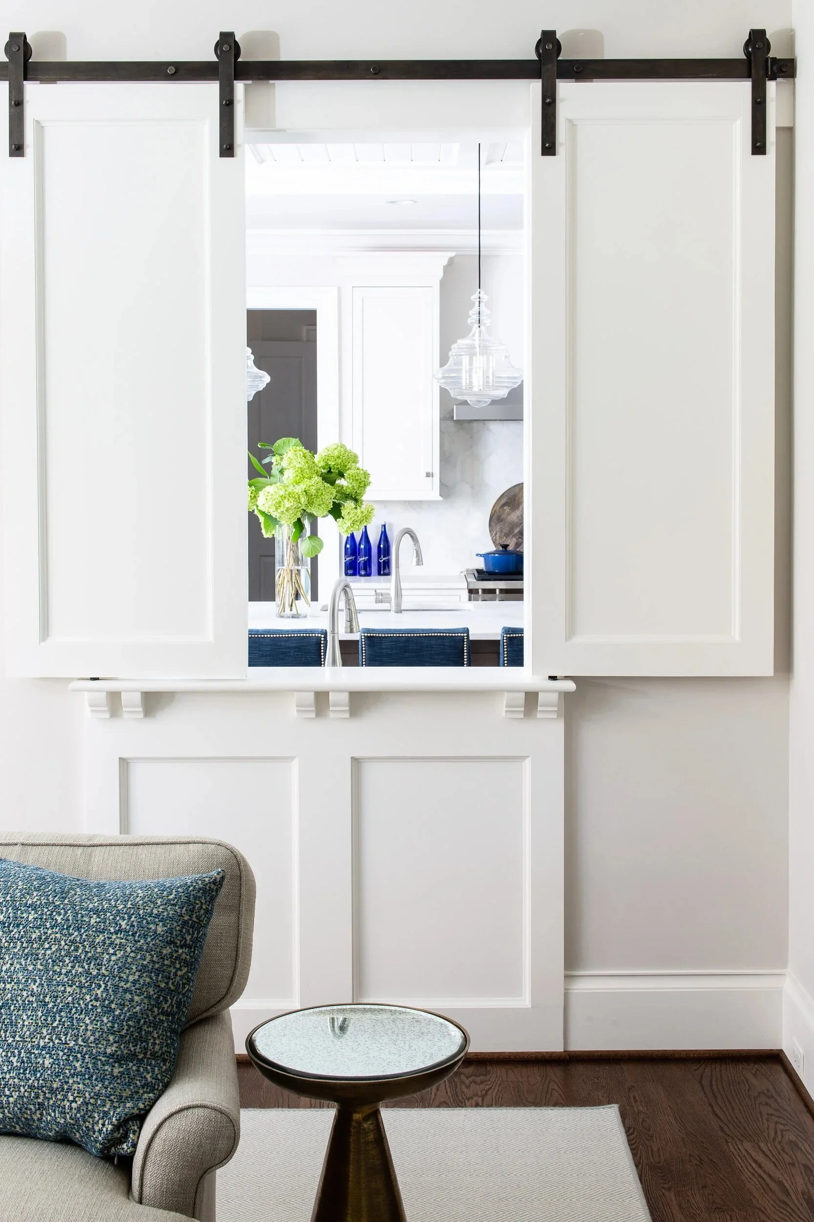View through a white sliding barn door to a kitchen with white cabinetry, blue vases, a vase with green hydrangeas, and pendant lighting.