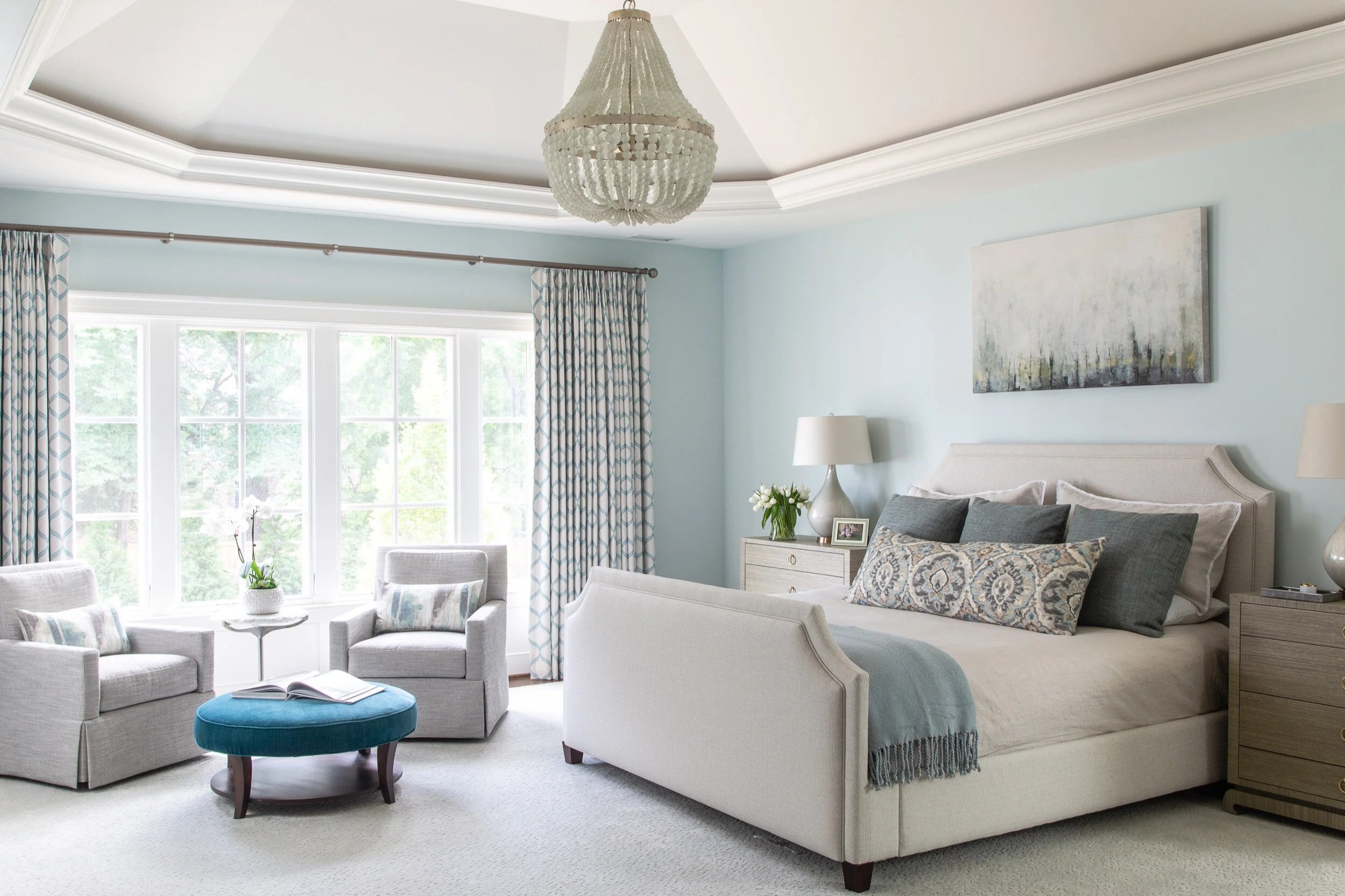 Bright bedroom with large window, white upholstered bed with multiple pillows, nightstands with lamps, a seating area with two armchairs, a blue ottoman with an open book, and a chandelier overhead.