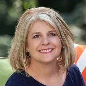 A woman with blonde hair smiling outdoors with greenery in the background.