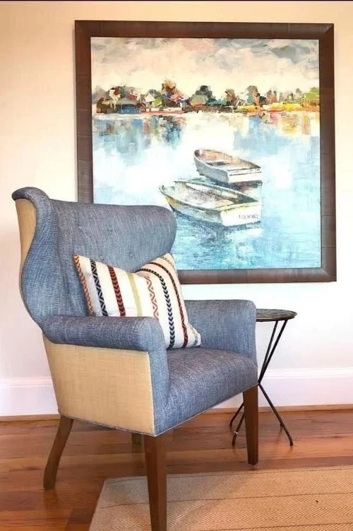 A cozy living corner featuring a high-back gray and beige armchair with a decorative striped pillow, a small black side table, and a large framed watercolor painting of boats on water hung on a white wall. Hardwood floor with a beige rug.