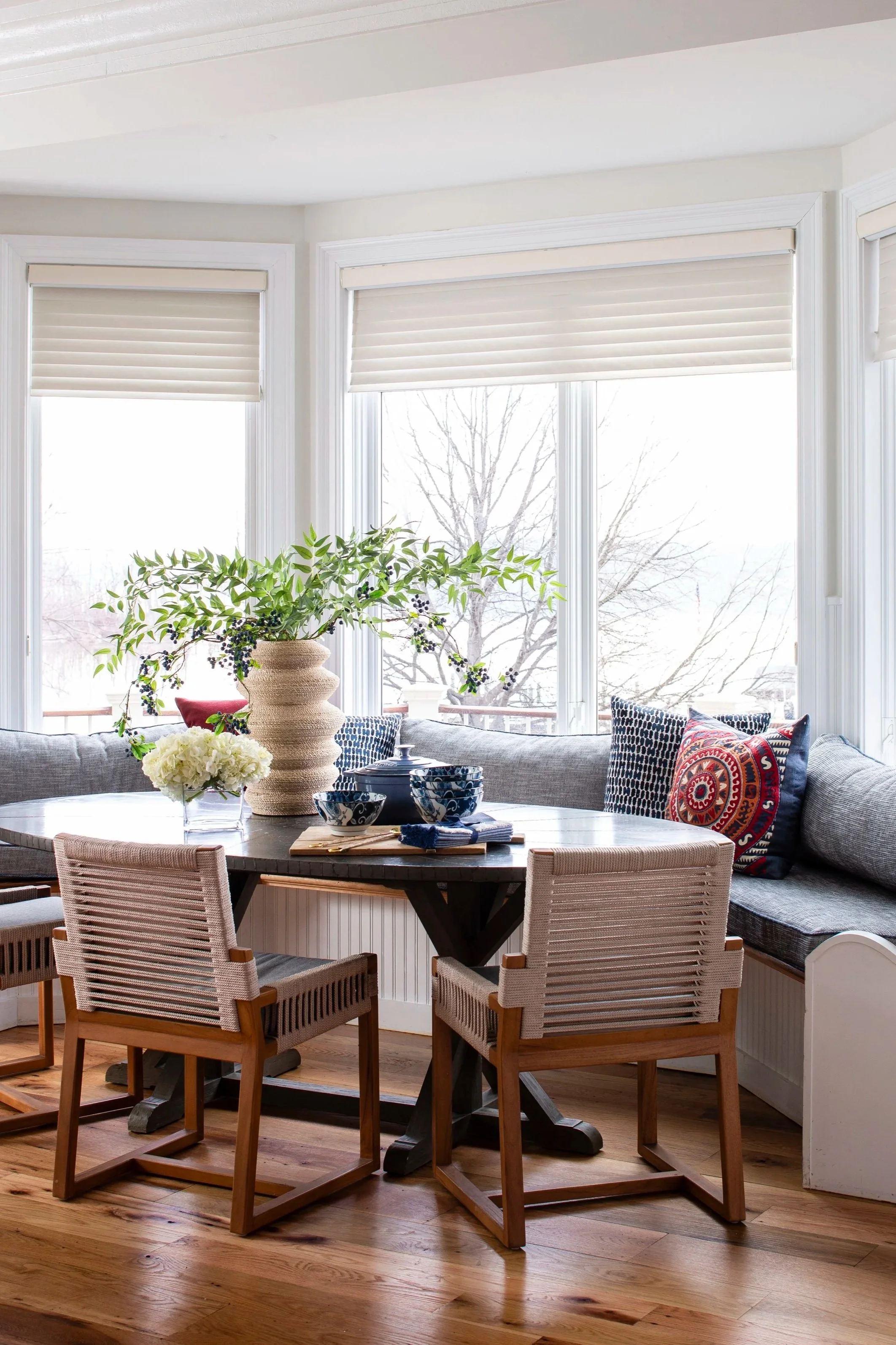 Dining area with a round wooden table, cushioned chairs, an extended window nook with cushions, a large flower vase, bowls, and a small potted plant, with large windows showing a snowy outdoor scene.