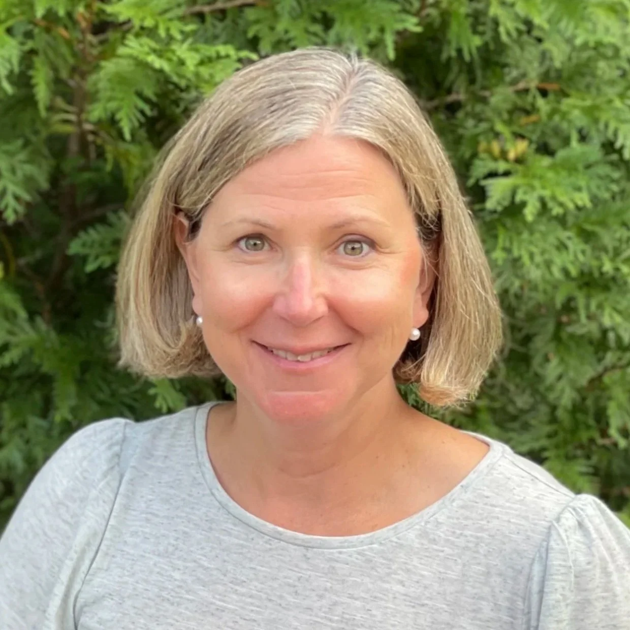 Close-up of a smiling woman with short blonde hair and wearing pearl earrings, standing outdoors with green foliage in the background.
