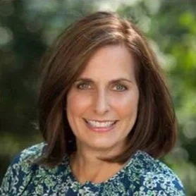A smiling woman with shoulder-length brown hair outdoors with greenery in the background.