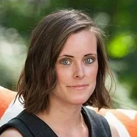 A woman with shoulder-length brown hair and blue eyes, looking directly at the camera, outdoors with green foliage in the background.