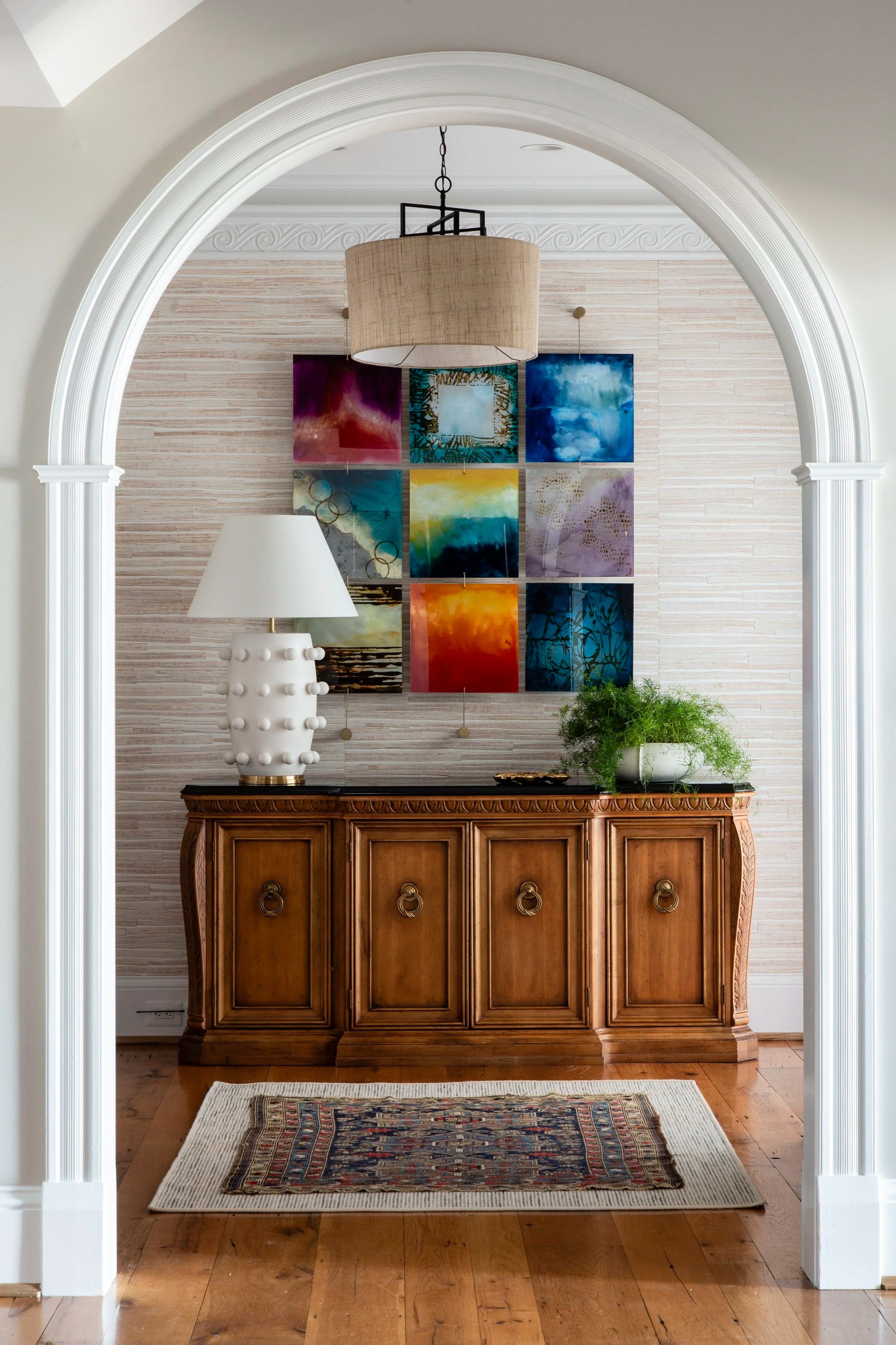 View through a white archway into a room with a wooden sideboard, a white ceramic lamp, a green potted plant, a colorful abstract art piece on the wall, and a hanging drum shade ceiling light.
