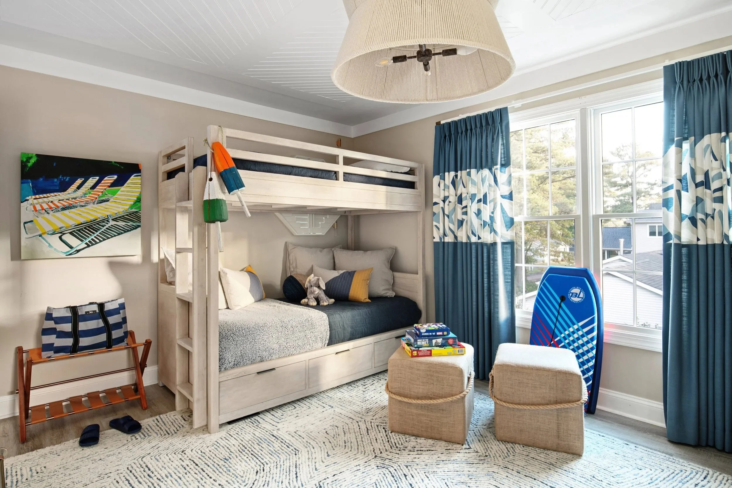 Child's bedroom with bunk beds, blue curtains, a surfboard, a painting, a stack of books, and a window showing trees and neighboring houses.