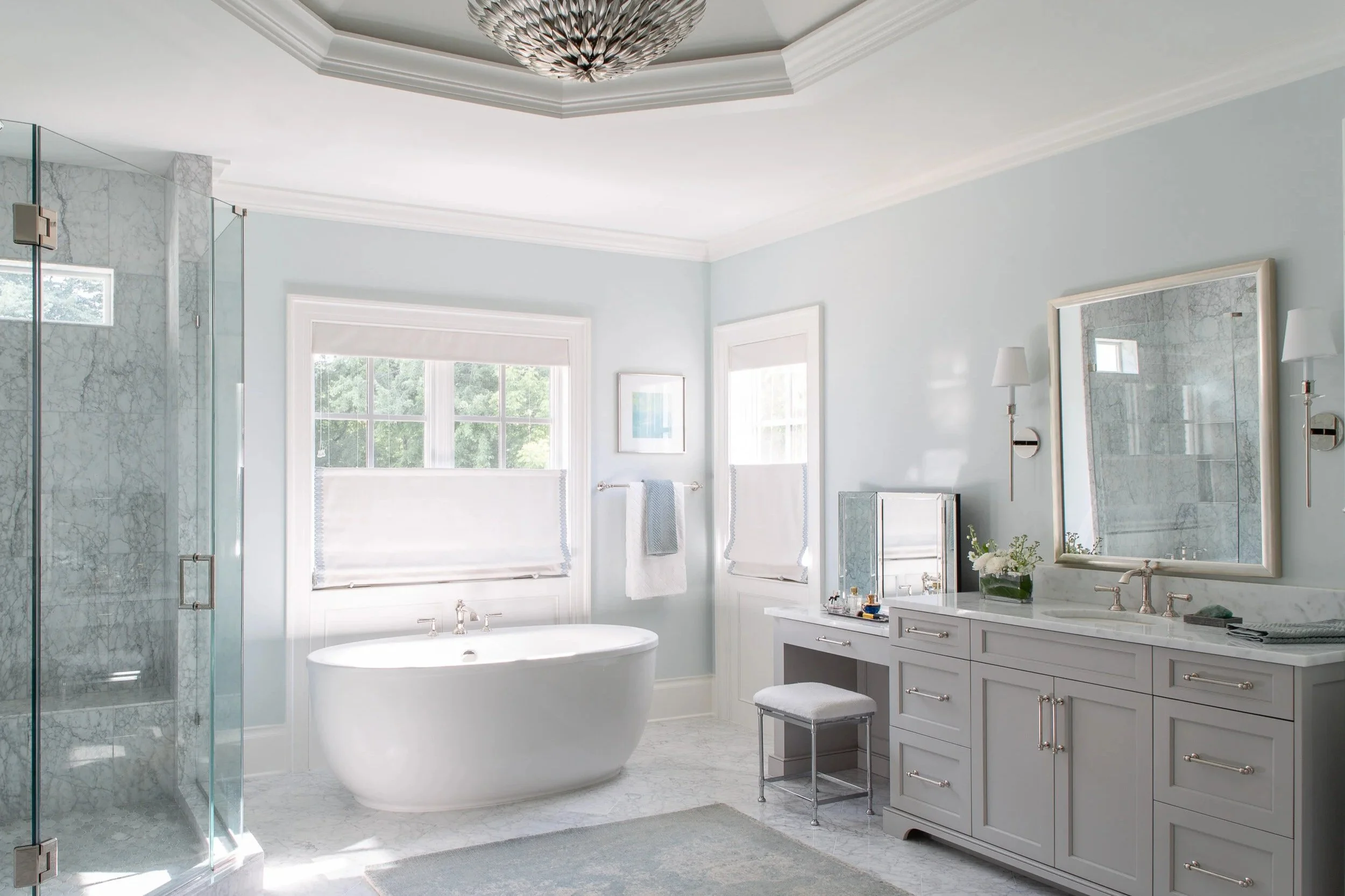 Bright and elegant bathroom with light blue walls, a large window with white blinds, a white bathtub beneath the window, a marble-topped vanity with a large mirror, modern wall sconces, a small seating stool, towel bars with white towels, a glass shower enclosure with marble interior, and decorative accessories.