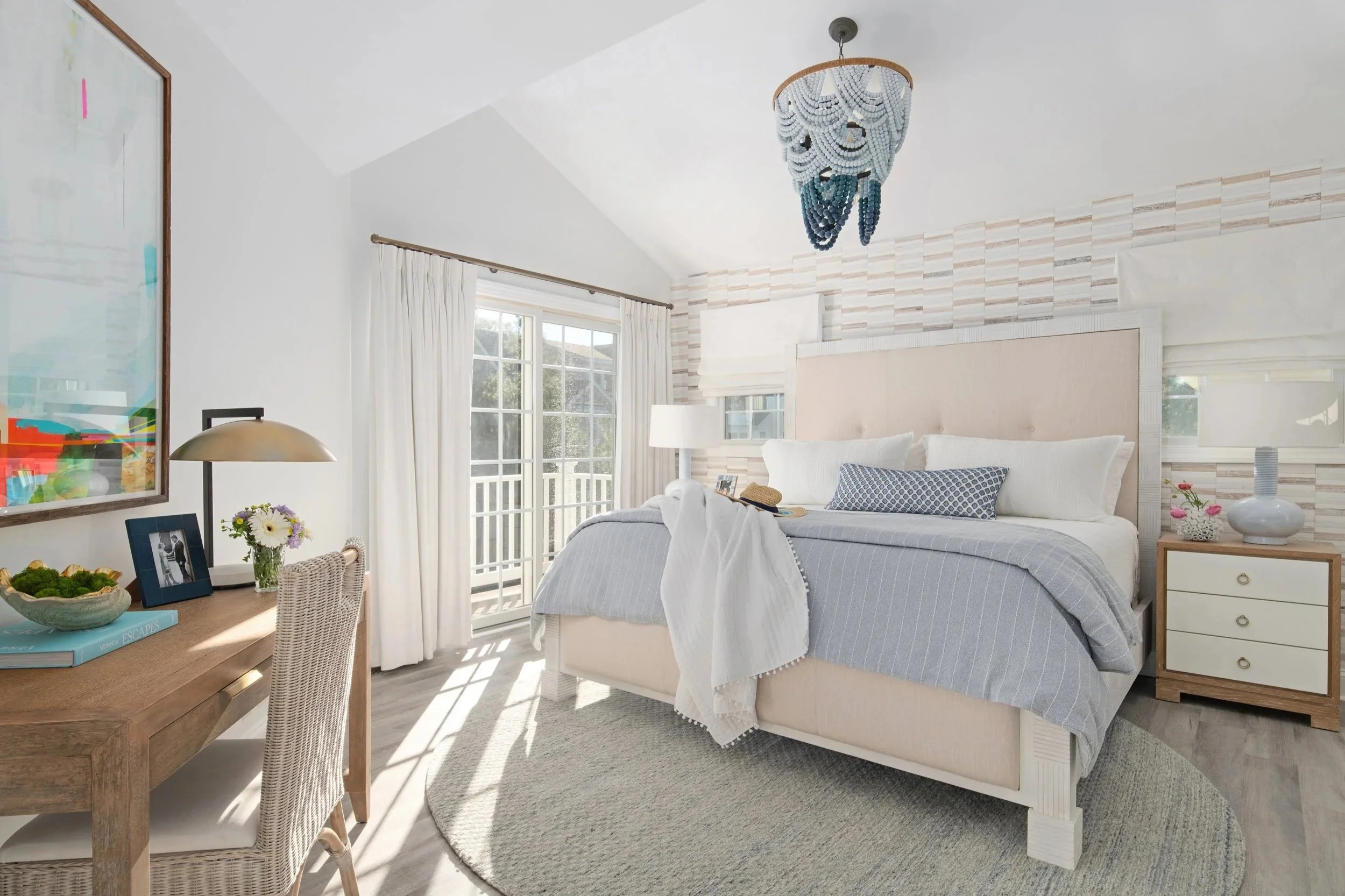 Cozy bedroom with a large bed, lamps, artwork, and a sliding glass door leading to a balcony, decorated in light tones with a beaded chandelier hanging overhead.