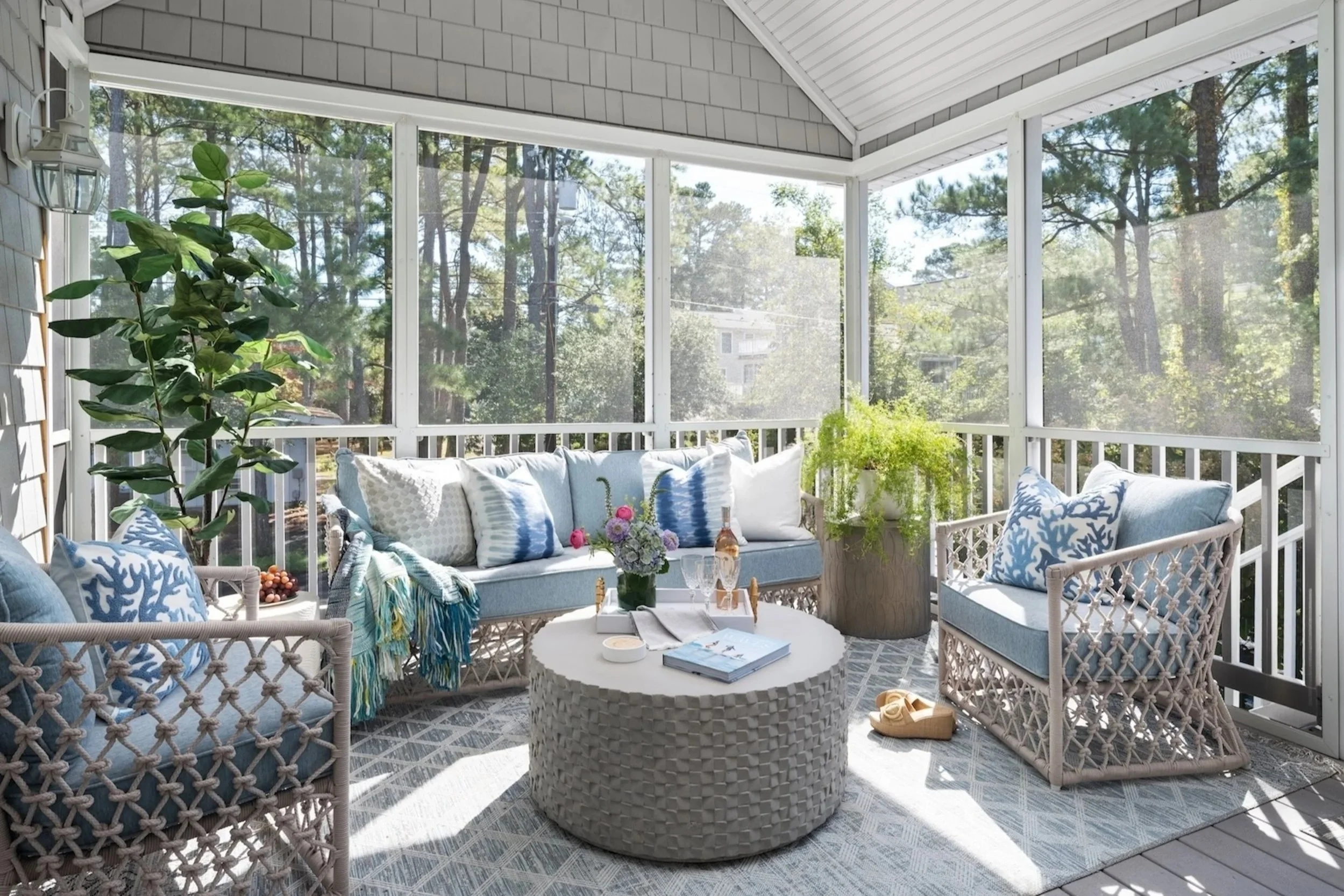 Sunlit screened porch with wicker furniture, cushions, throw blankets, potted plants, and a round table with a book, flowers, and drinks, overlooking trees.